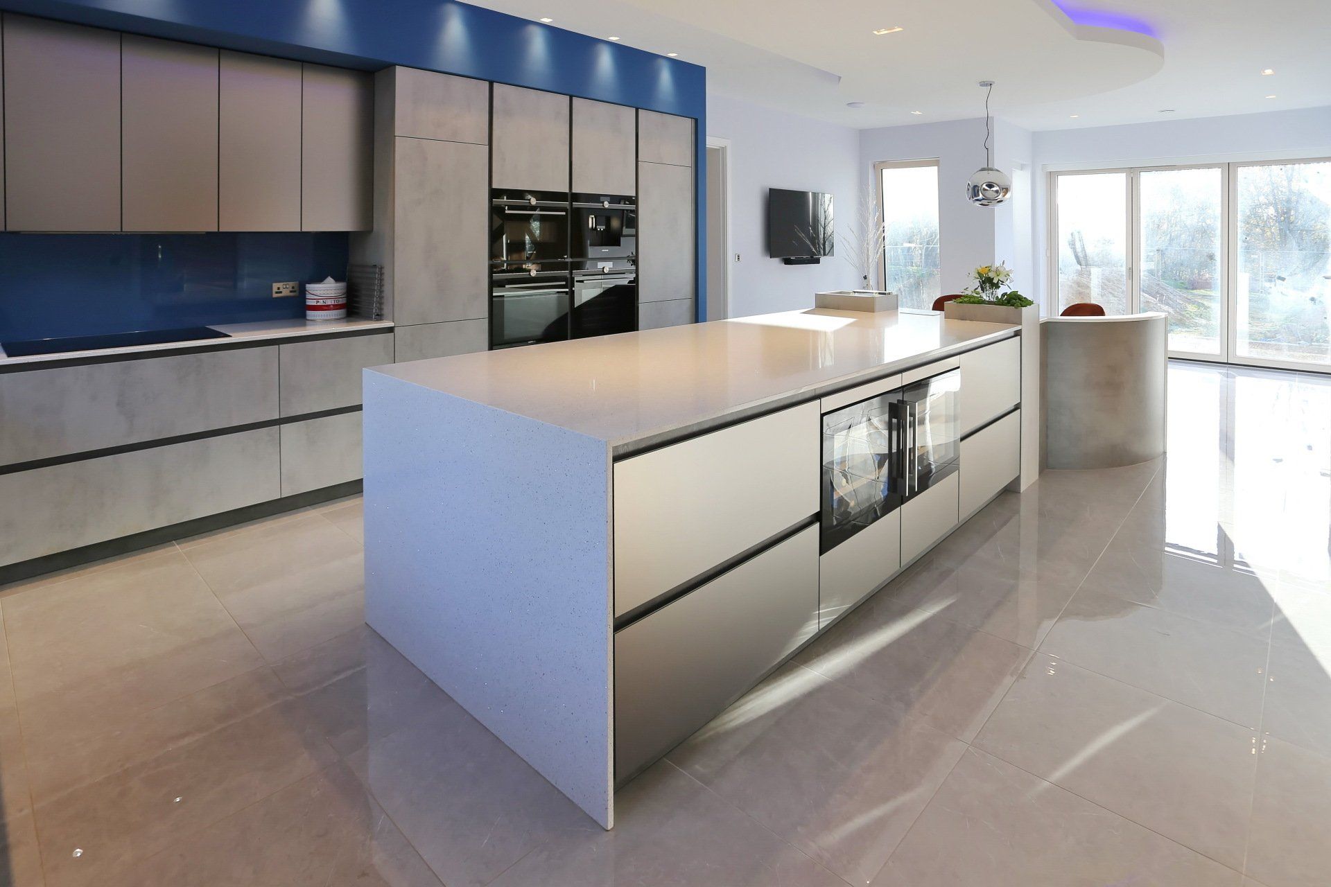 Modern kitchen with large island, stainless steel cabinets, blue backsplash, and light-colored flooring.