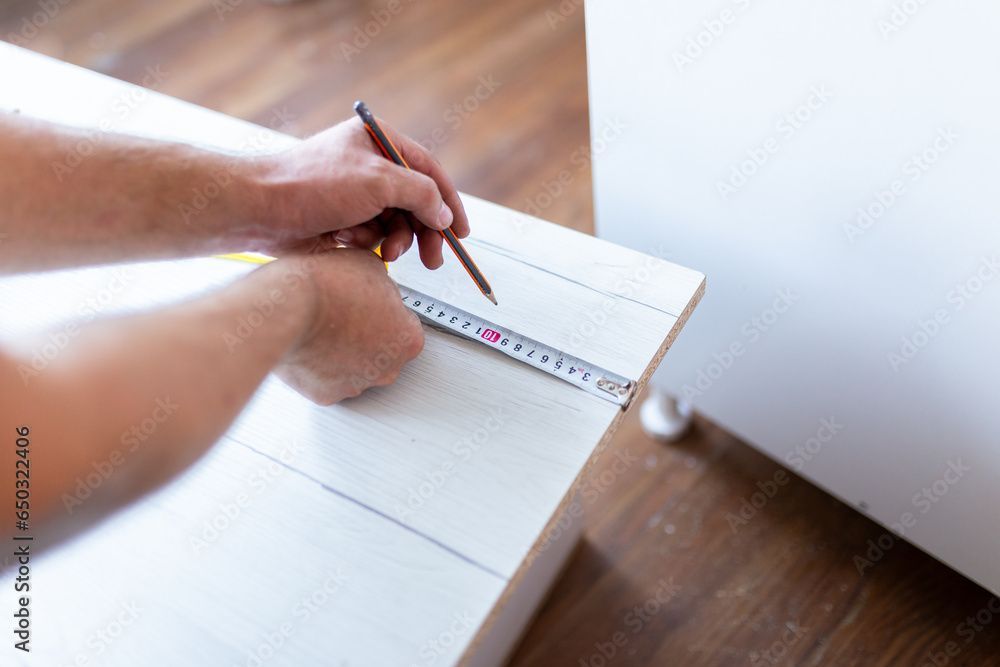 Person measures and marks wood with a pencil, using a tape measure.