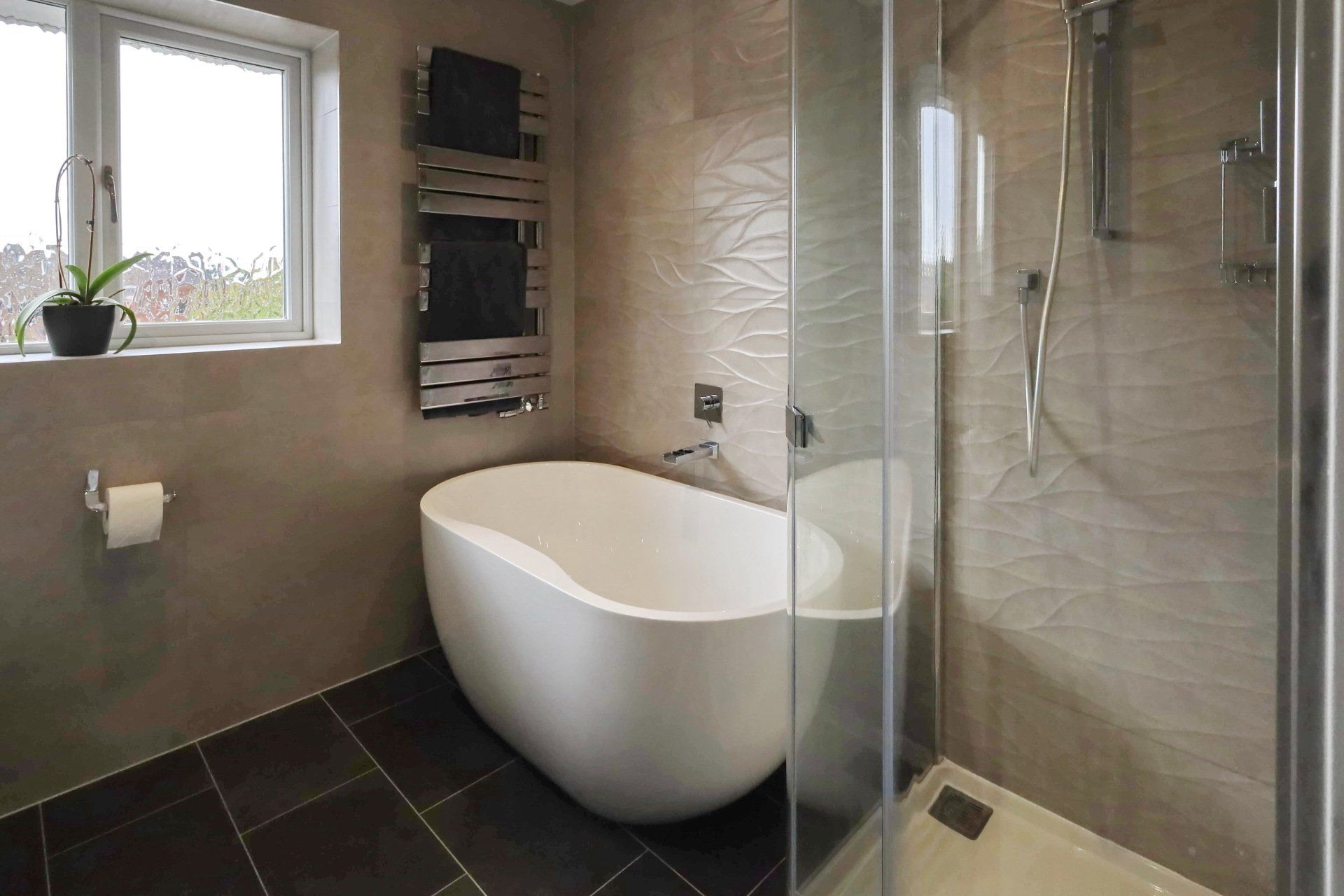 Modern bathroom with white tub, glass shower, towel rack, window, and dark tile floor.