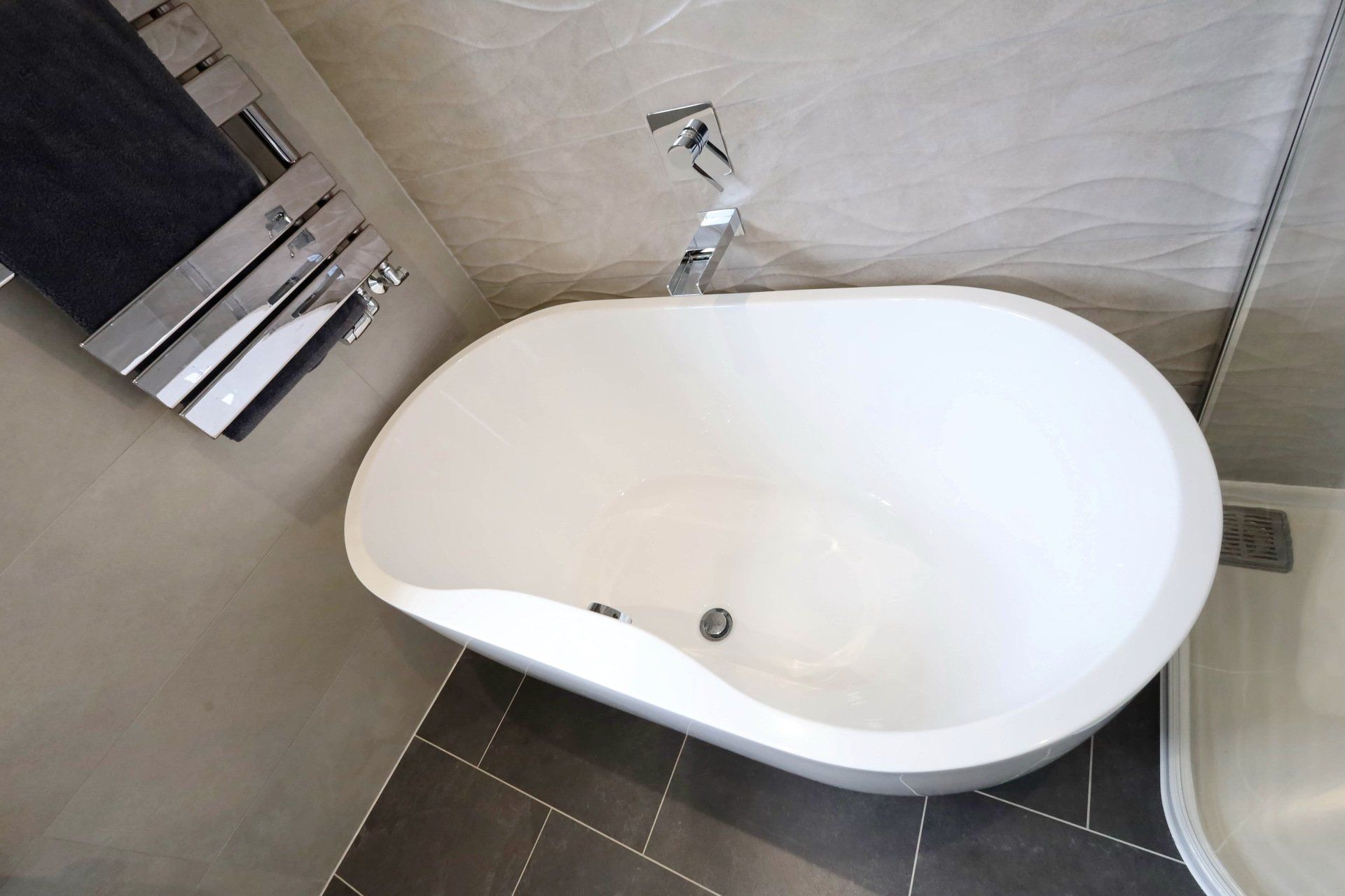 Oval-shaped white bathtub in a modern bathroom, next to a towel rack and faucet. Dark tile floor.