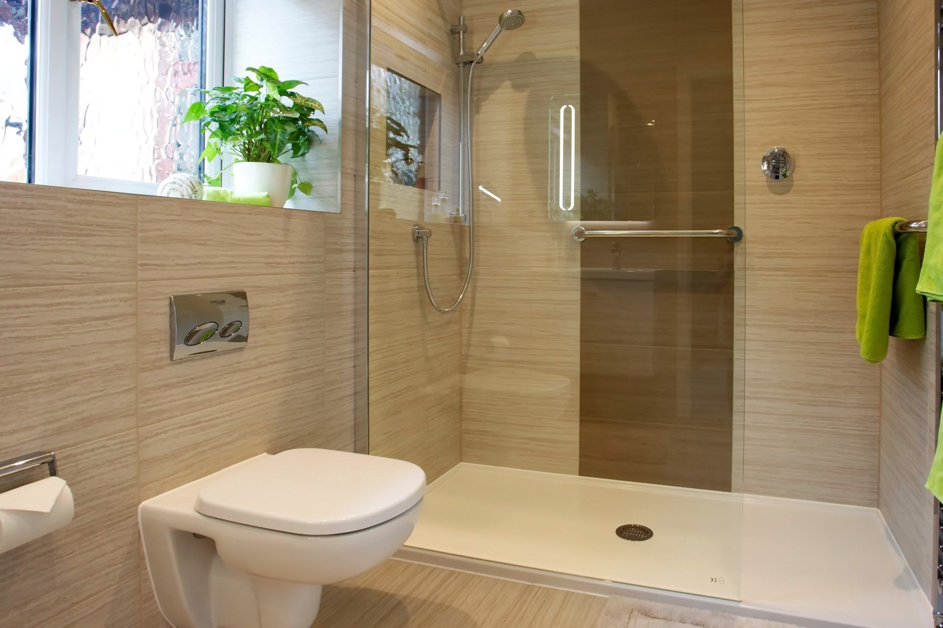 Modern bathroom with walk-in shower, wall-mounted toilet, and light-colored tiles. Green towels hang.