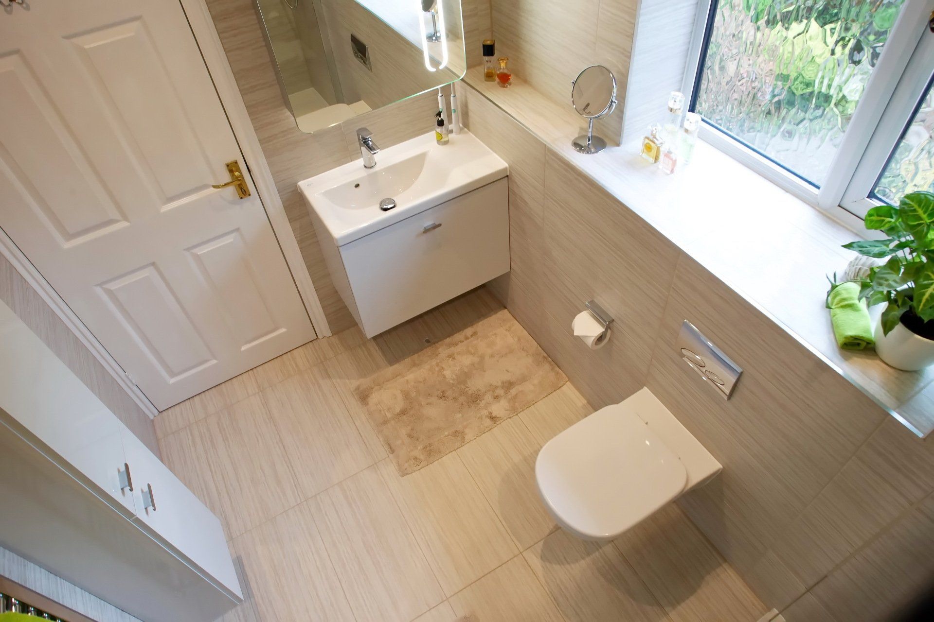 Small, modern bathroom with a white toilet, sink, and door. Natural light from a window with a green plant.