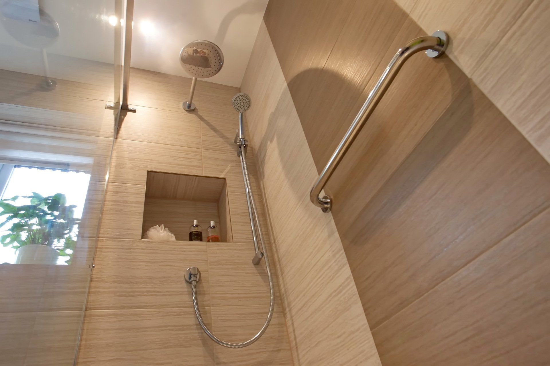 Shower with beige tiled walls, a handheld showerhead, grab bar, and recessed shelf.
