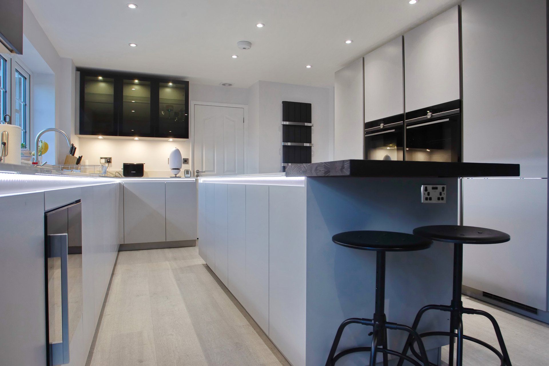 Modern white kitchen with island and black stools. Built-in appliances, recessed lighting, and wooden floor.