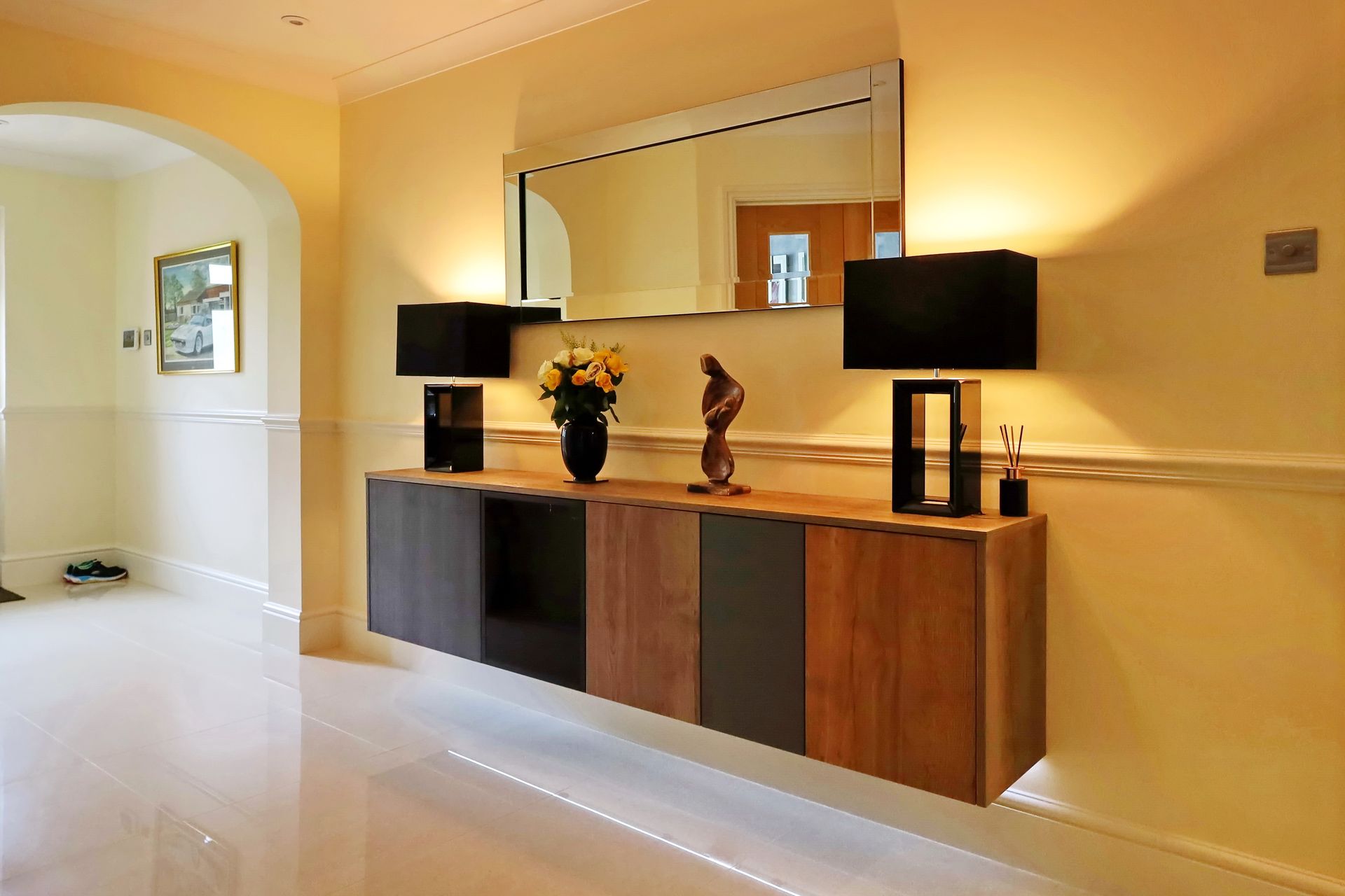Floating wood and gray cabinet with lamps, mirror, and flowers in a hallway.