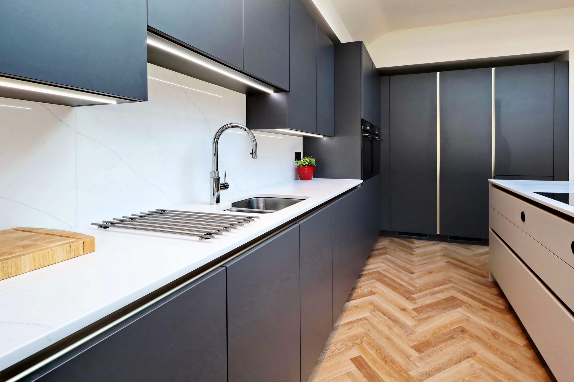 Modern kitchen with dark gray cabinets, white countertops, stainless steel sink, and wooden herringbone floor.