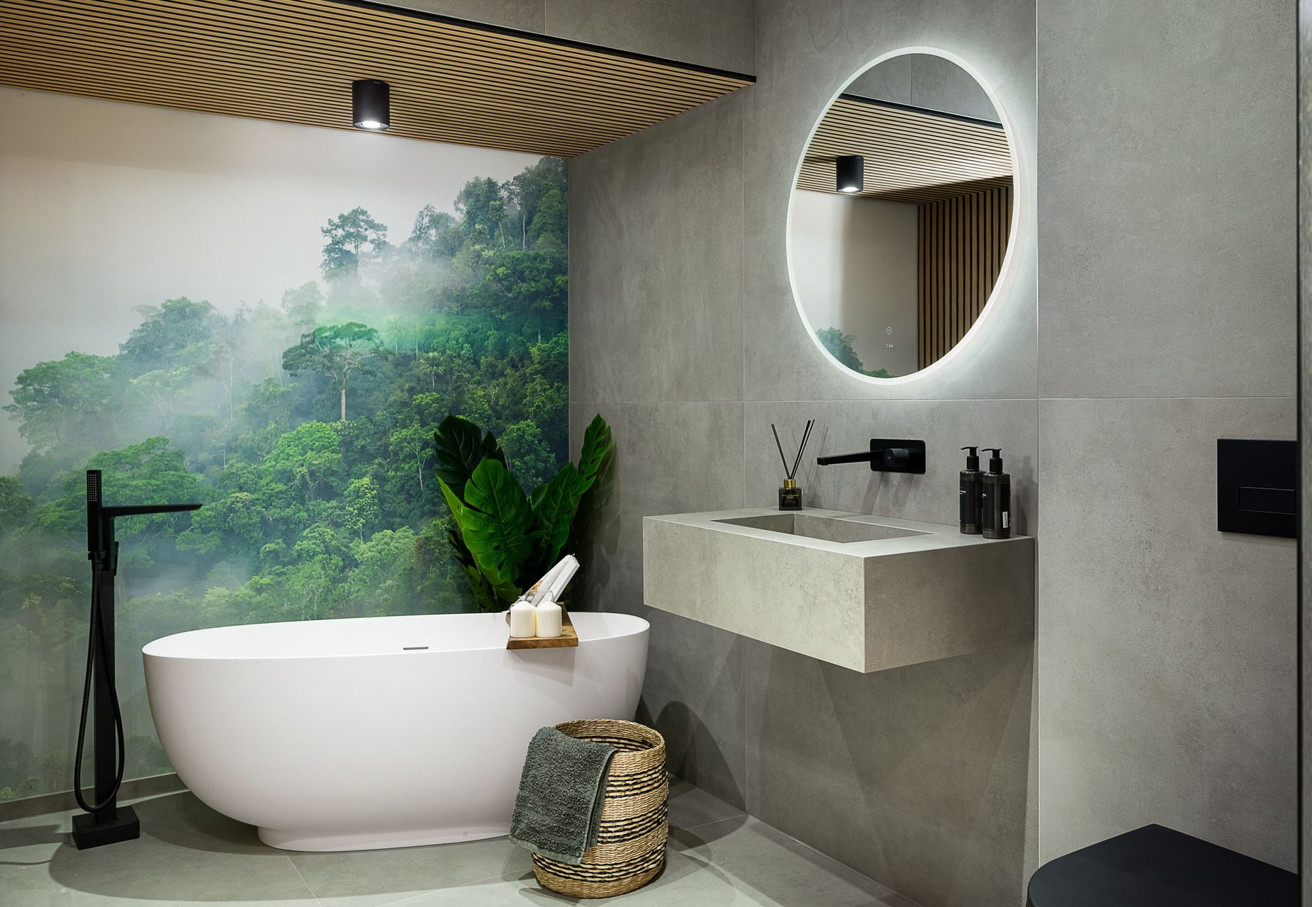 Modern bathroom with white tub, round mirror, concrete sink, forest mural.