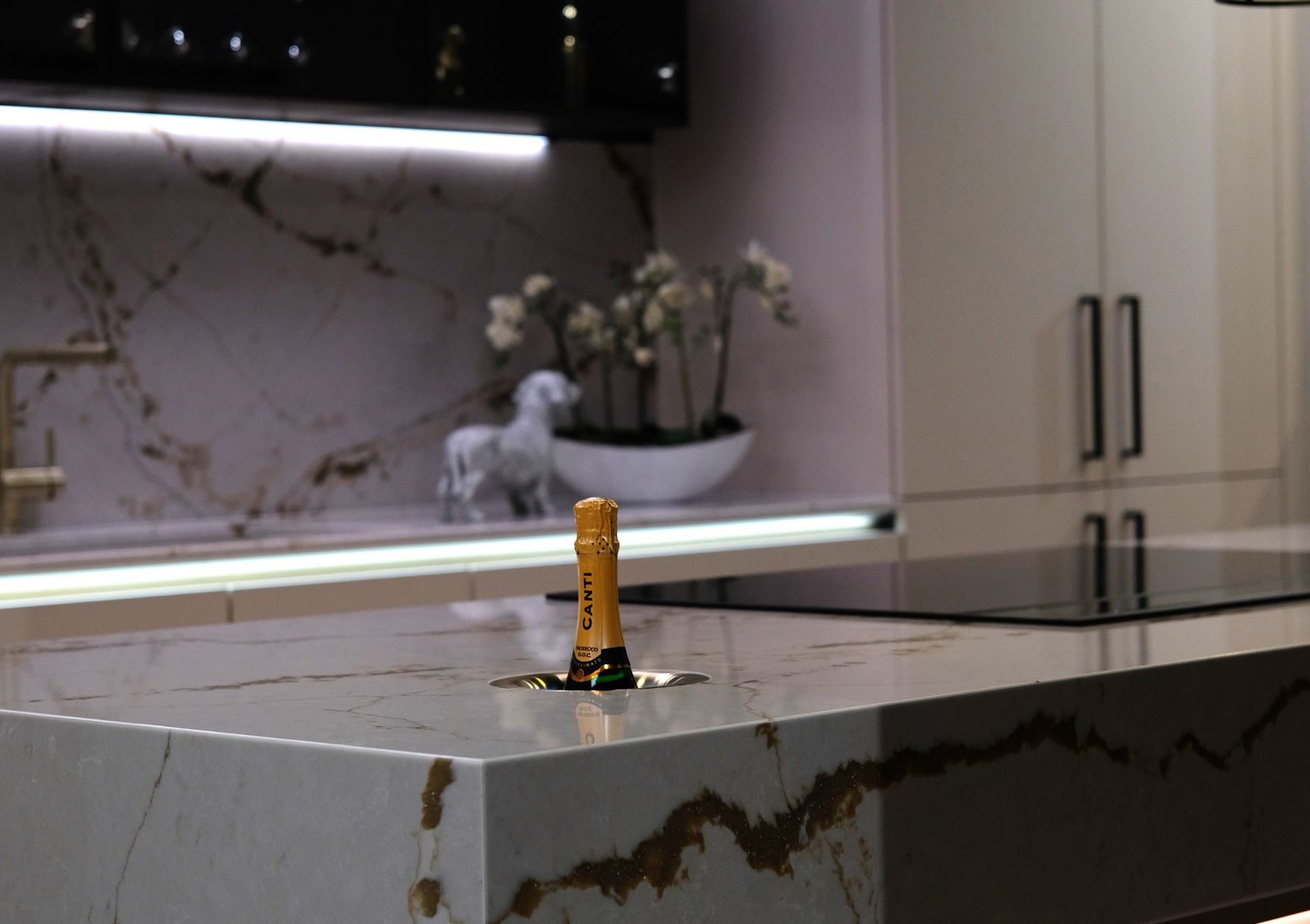A champagne bottle sits in a kitchen countertop. The countertop is marble with gold veining, and cabinets are visible.