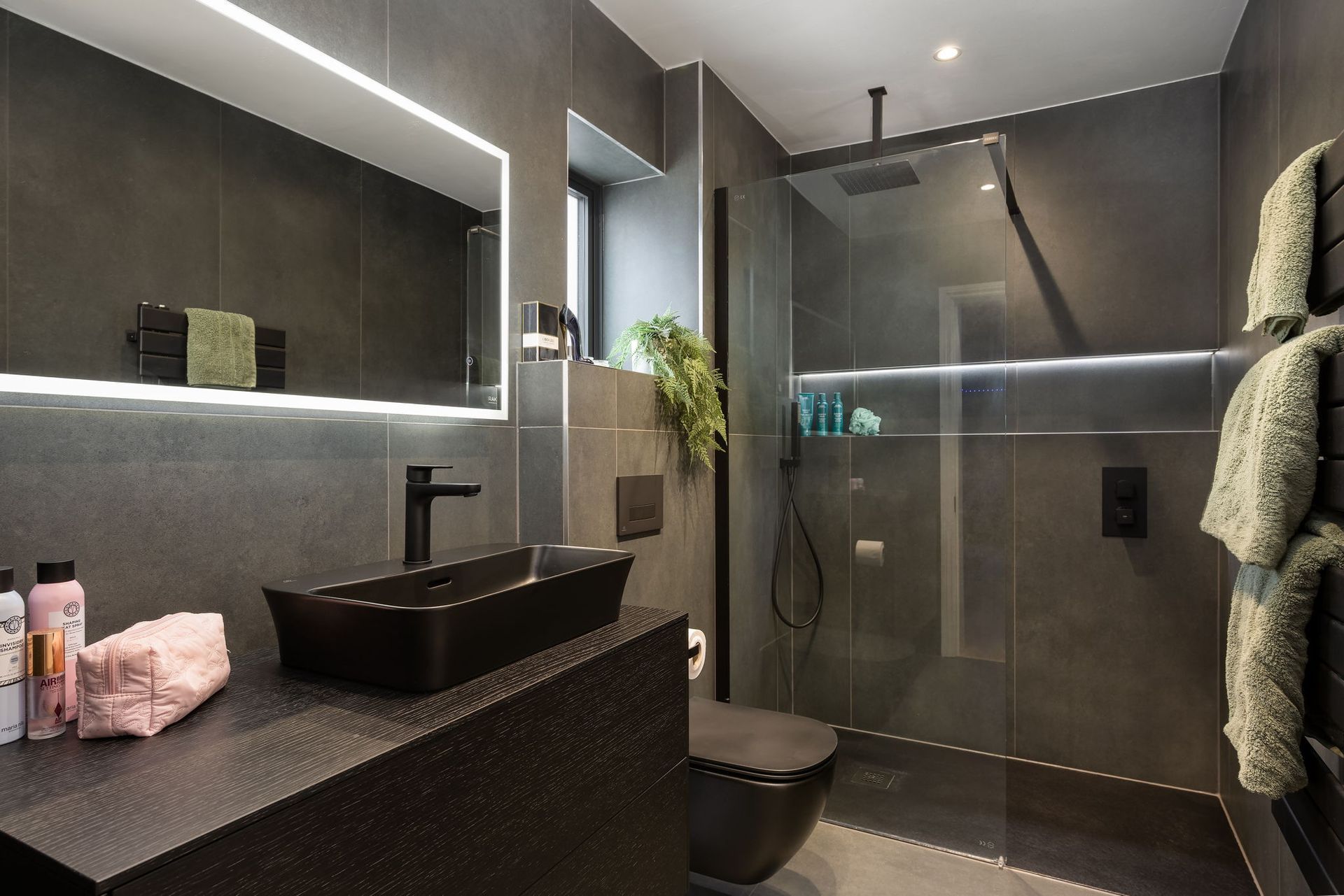 Modern, dark gray bathroom with black sink, shower, and toilet. Features a lit mirror and shelves.