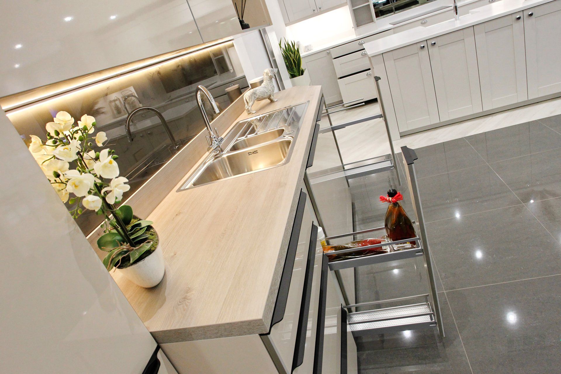 Modern kitchen with light wood countertop and stainless steel sink. Pull-out shelving with jars and bottles.