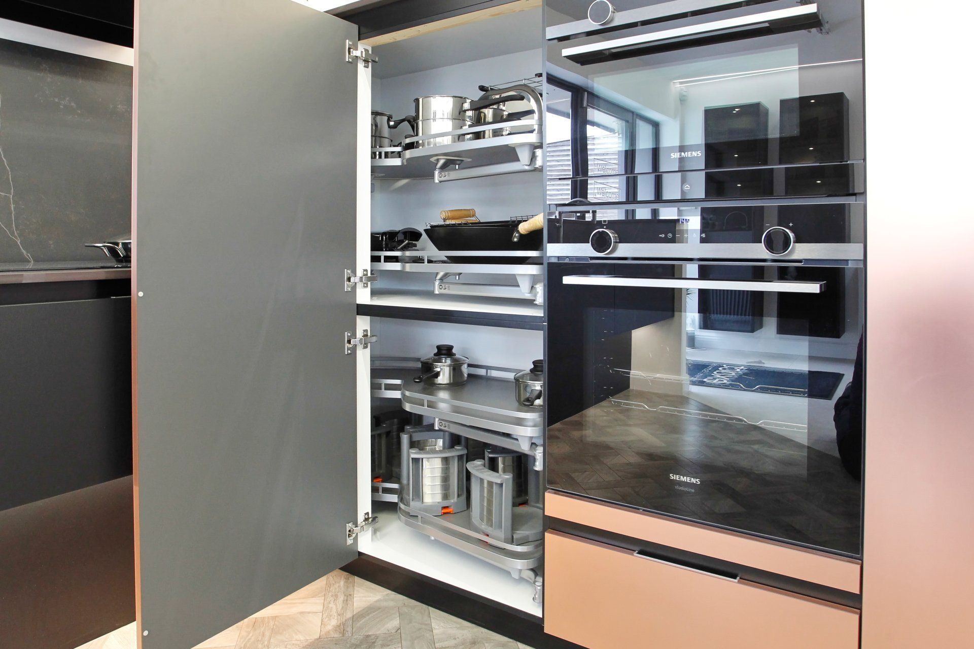 Open kitchen cabinet with pull-out shelves holding cookware. Next to built-in ovens and rose gold countertop.