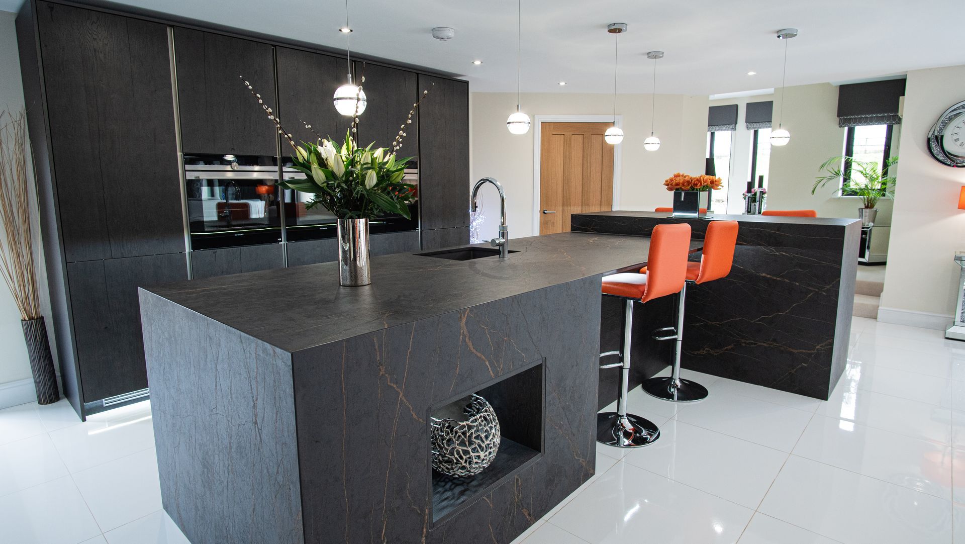 Modern kitchen with dark grey cabinetry and island, orange bar stools, white floor.