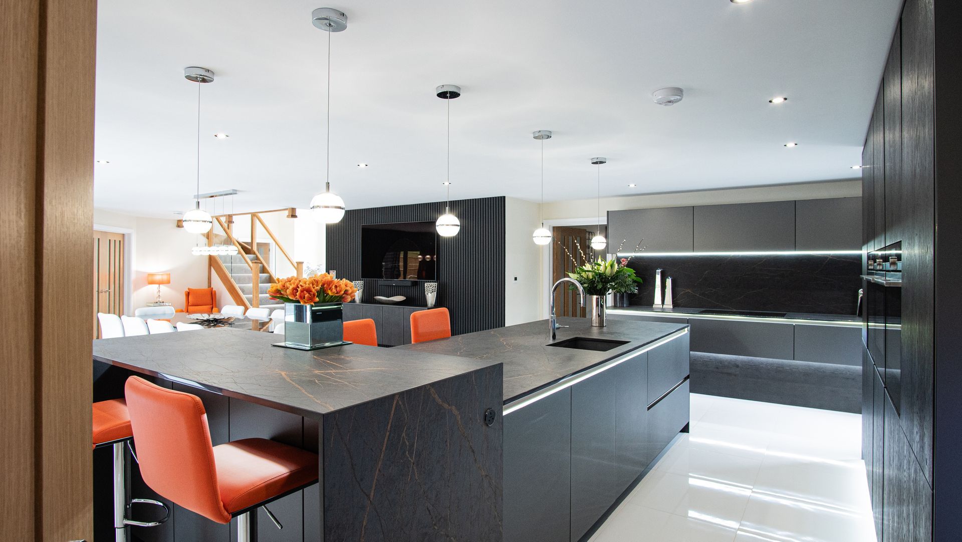 Modern kitchen with black cabinets, large island, orange stools, and pendant lights.