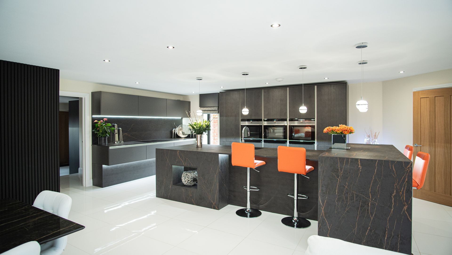 Modern kitchen with dark cabinetry, large island, orange bar stools, and white tile floor.