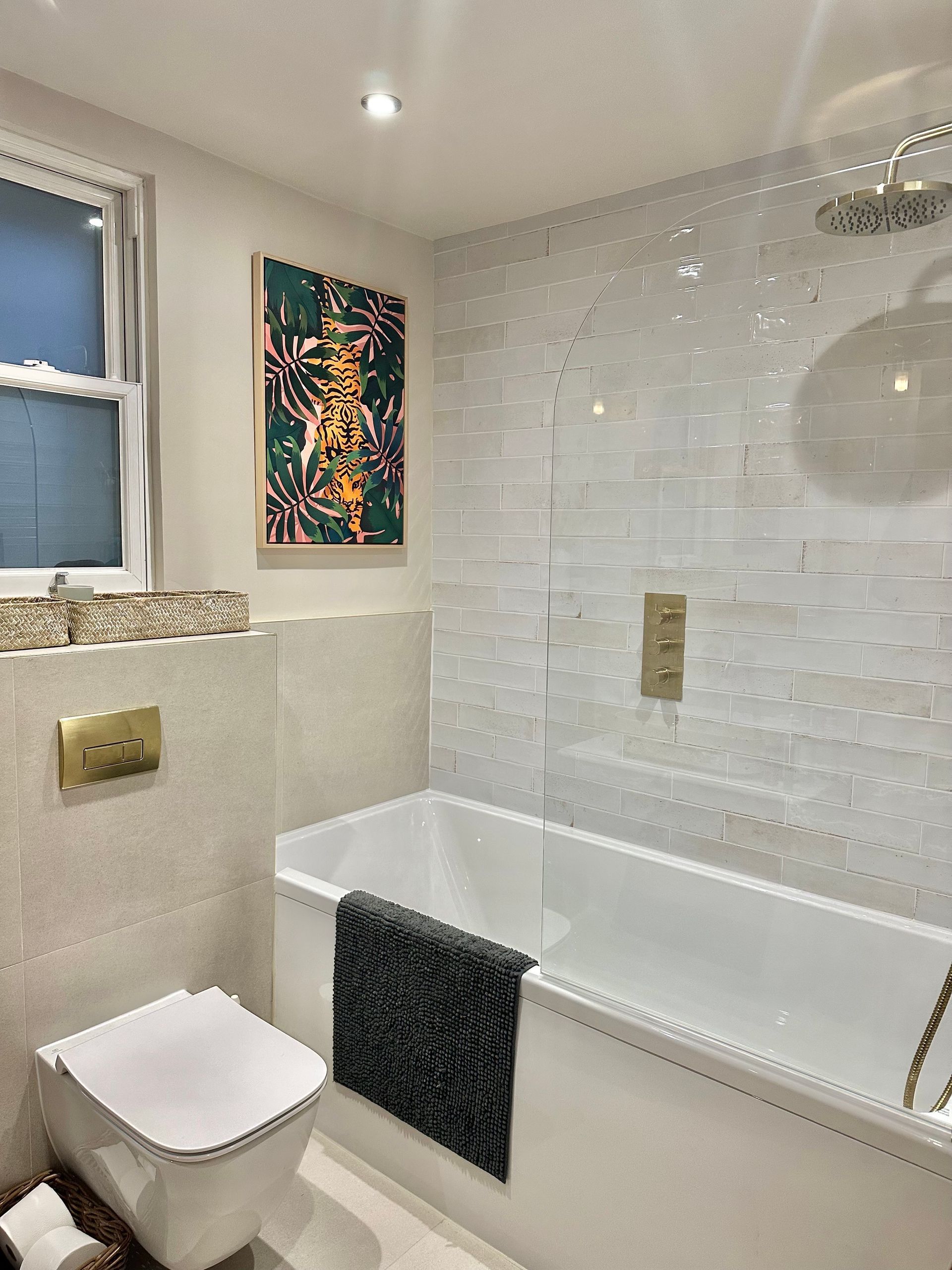 Modern bathroom with white tub, toilet, and mosaic tile walls; gold fixtures, art on wall.