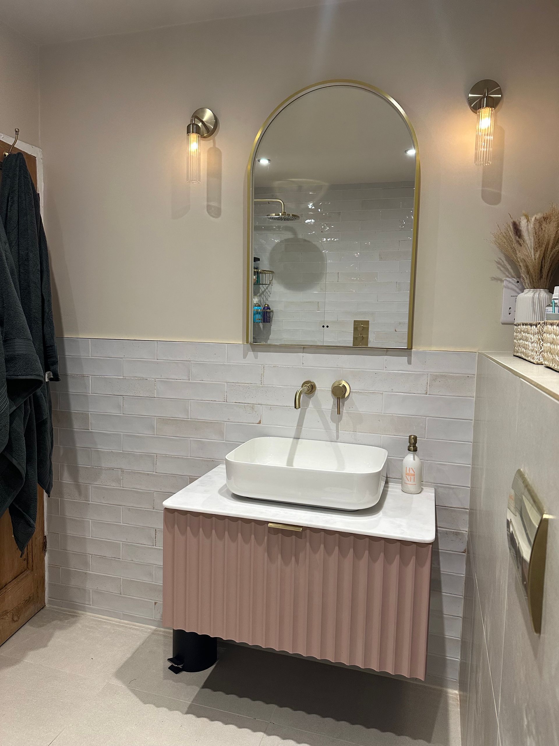 Modern bathroom with pink vanity, gold fixtures, and arched mirror.