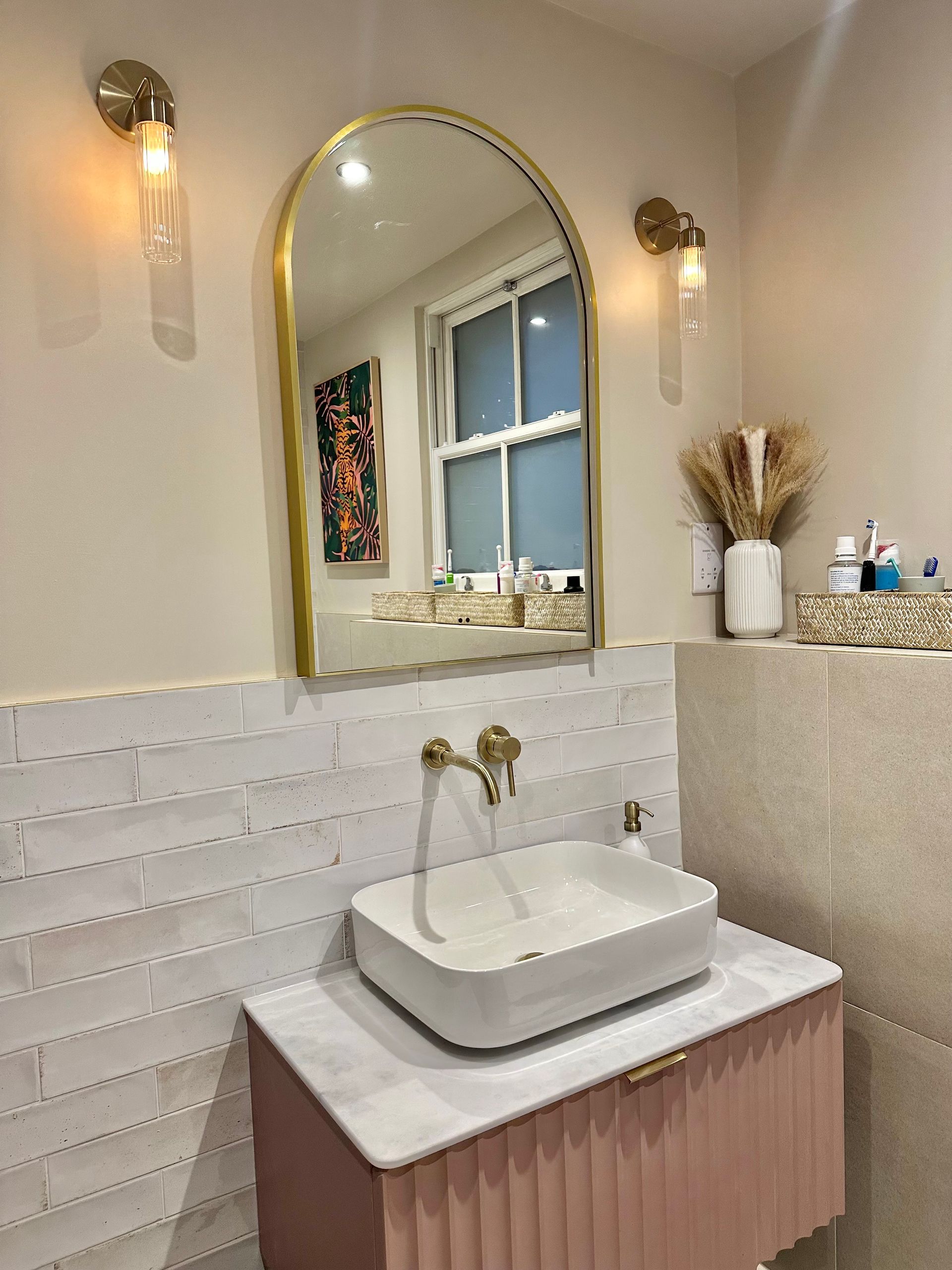 Bathroom with gold-framed arch mirror, white sink, pink cabinet, and decorative wall sconces.