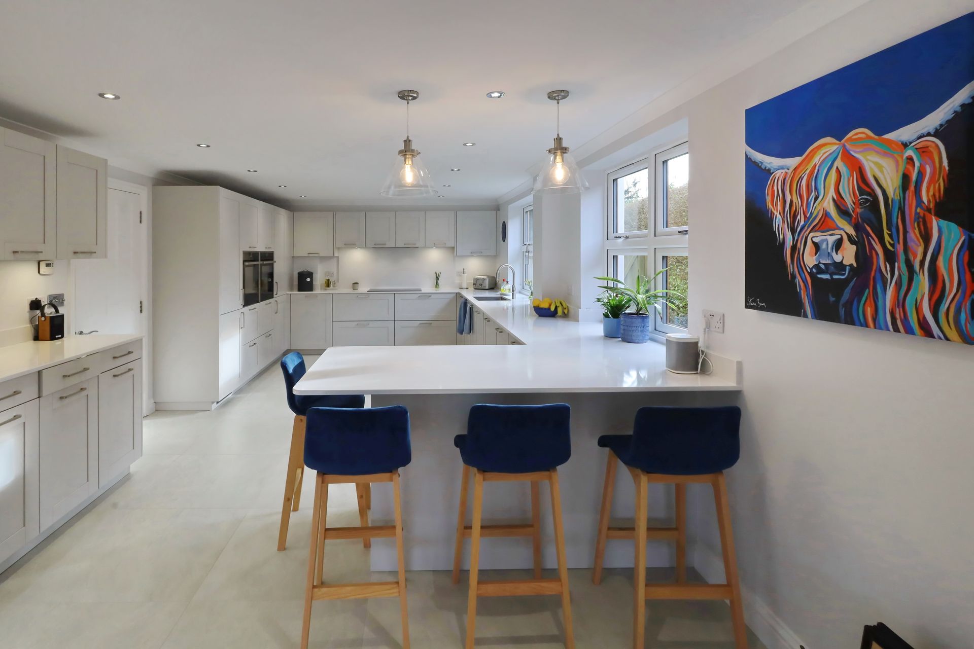 Modern kitchen with white cabinets, breakfast bar with blue stools, and a colorful cow painting.