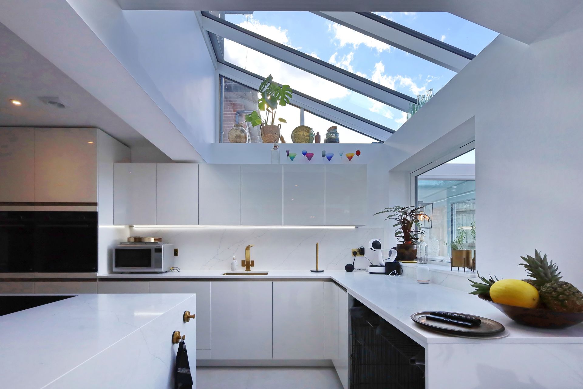 Modern white kitchen with angled skylights, counter, and cabinets. Sunlight illuminates the space.