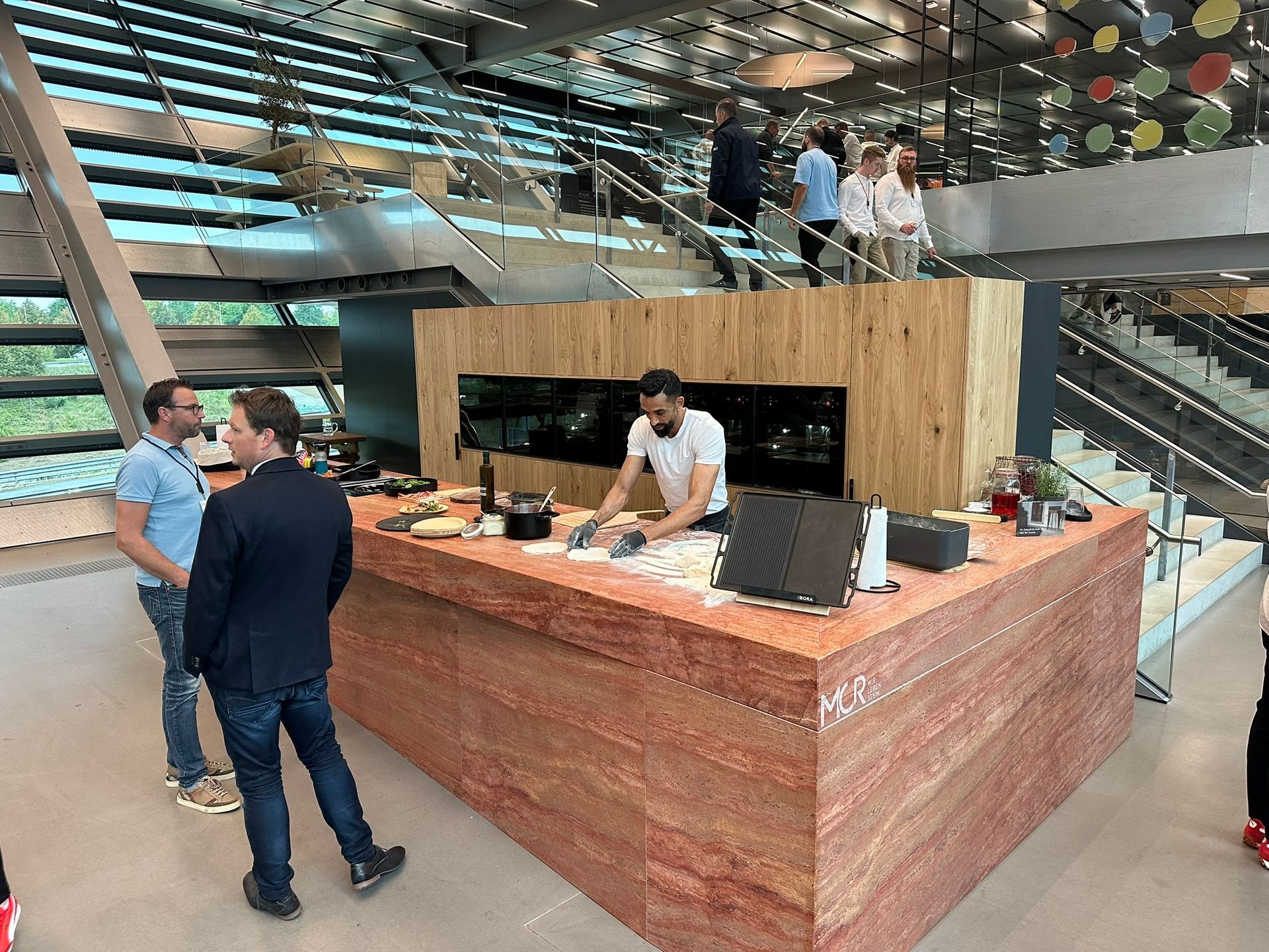 Chef preparing food at an island counter, with two men observing. Modern interior with stairs.