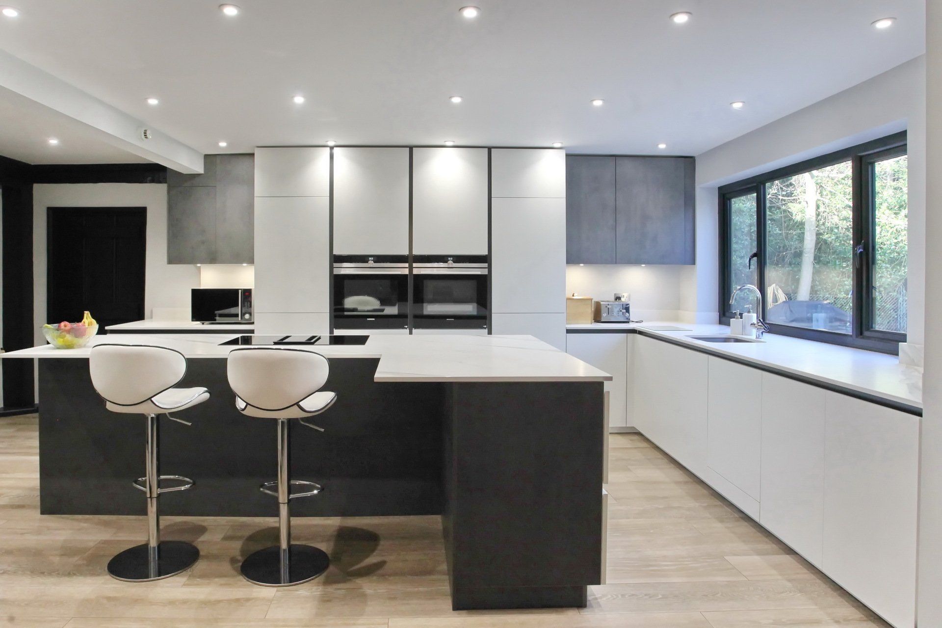 Modern kitchen with gray and white cabinetry, island with bar stools, window, and recessed lighting.