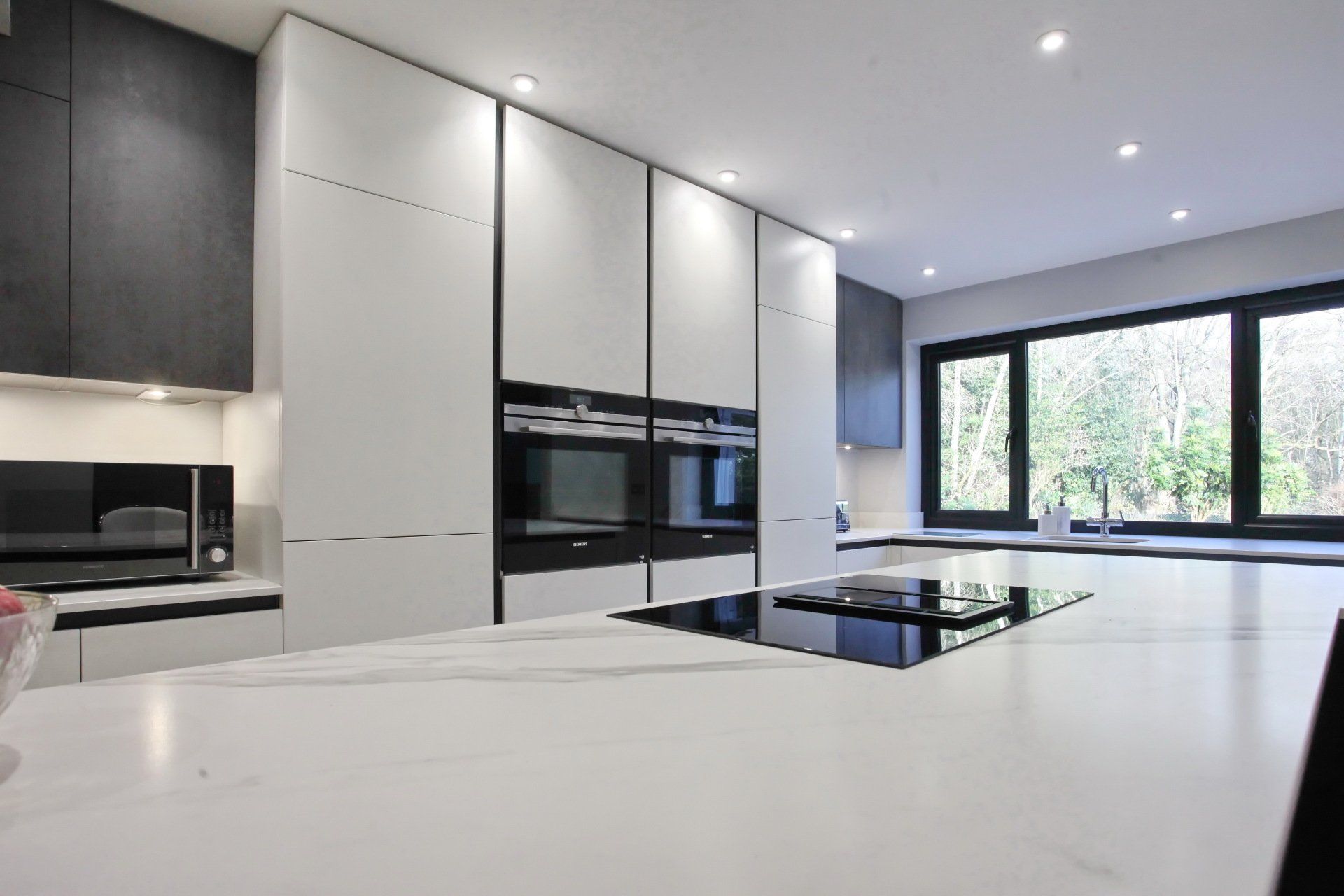 Modern kitchen with white countertops, black appliances, large windows, and overhead lighting.