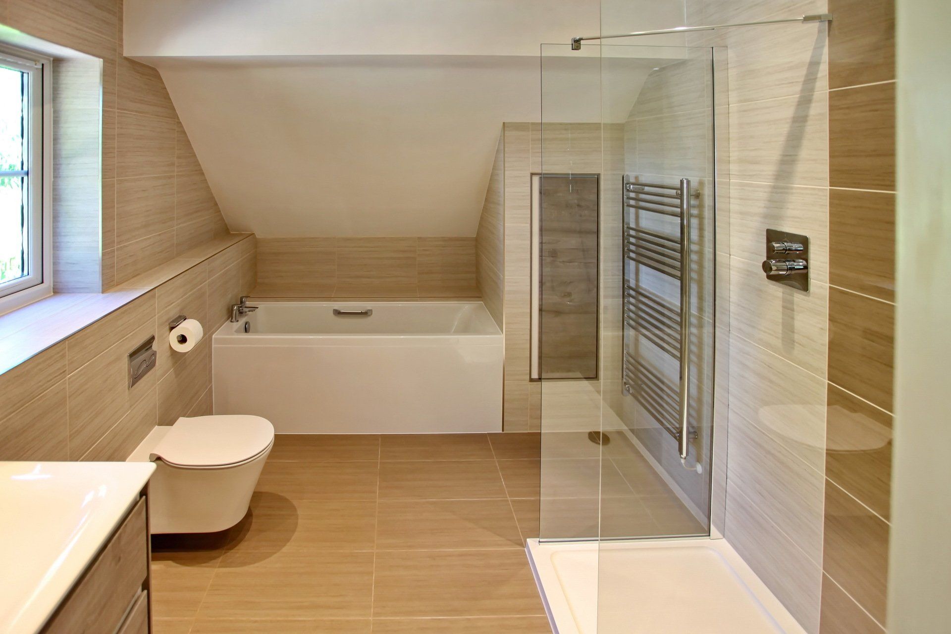 Modern bathroom with tan tile, shower, bathtub, and toilet under a sloped ceiling.