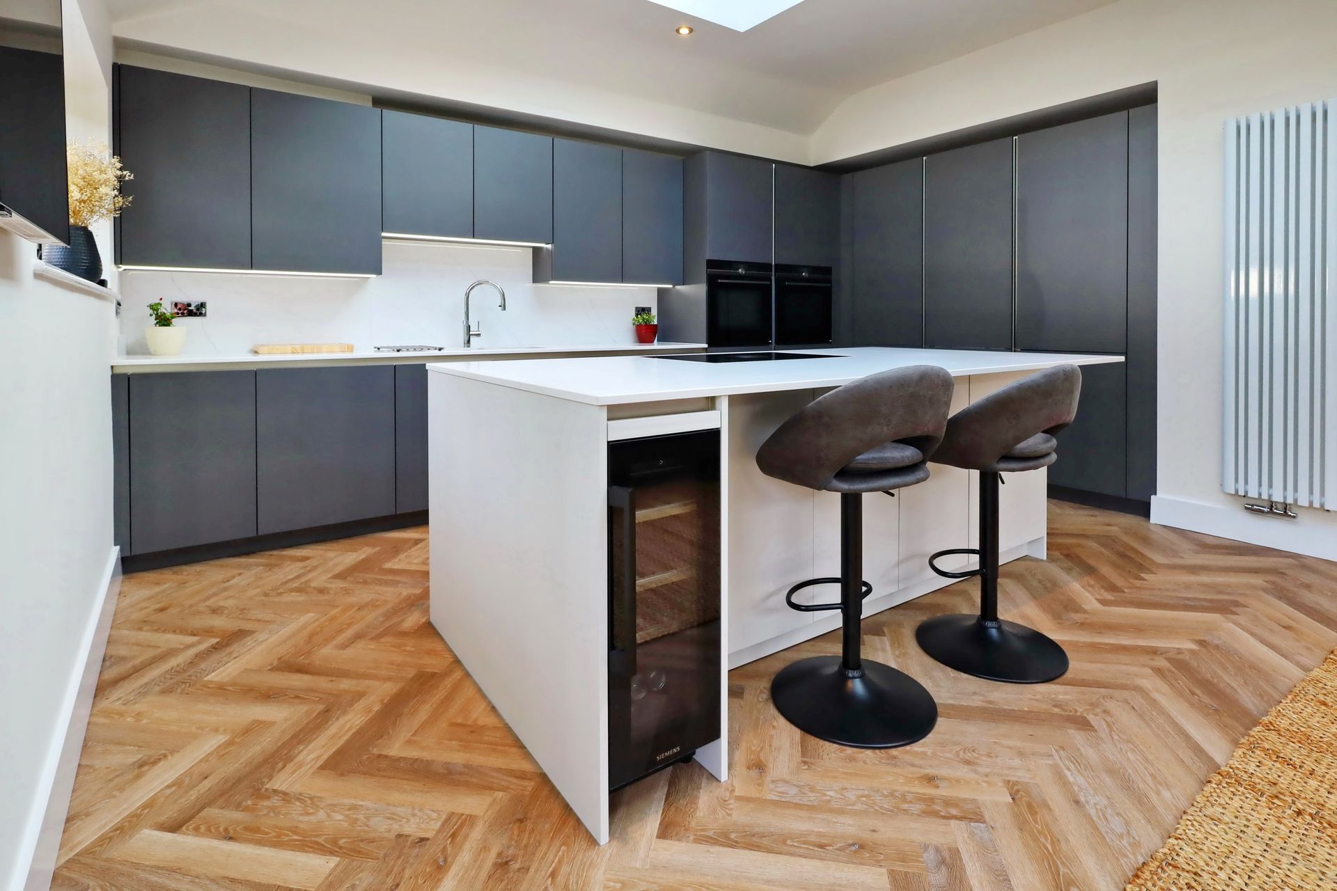 Modern kitchen with gray cabinets, white island, two bar stools, and herringbone wood floor.