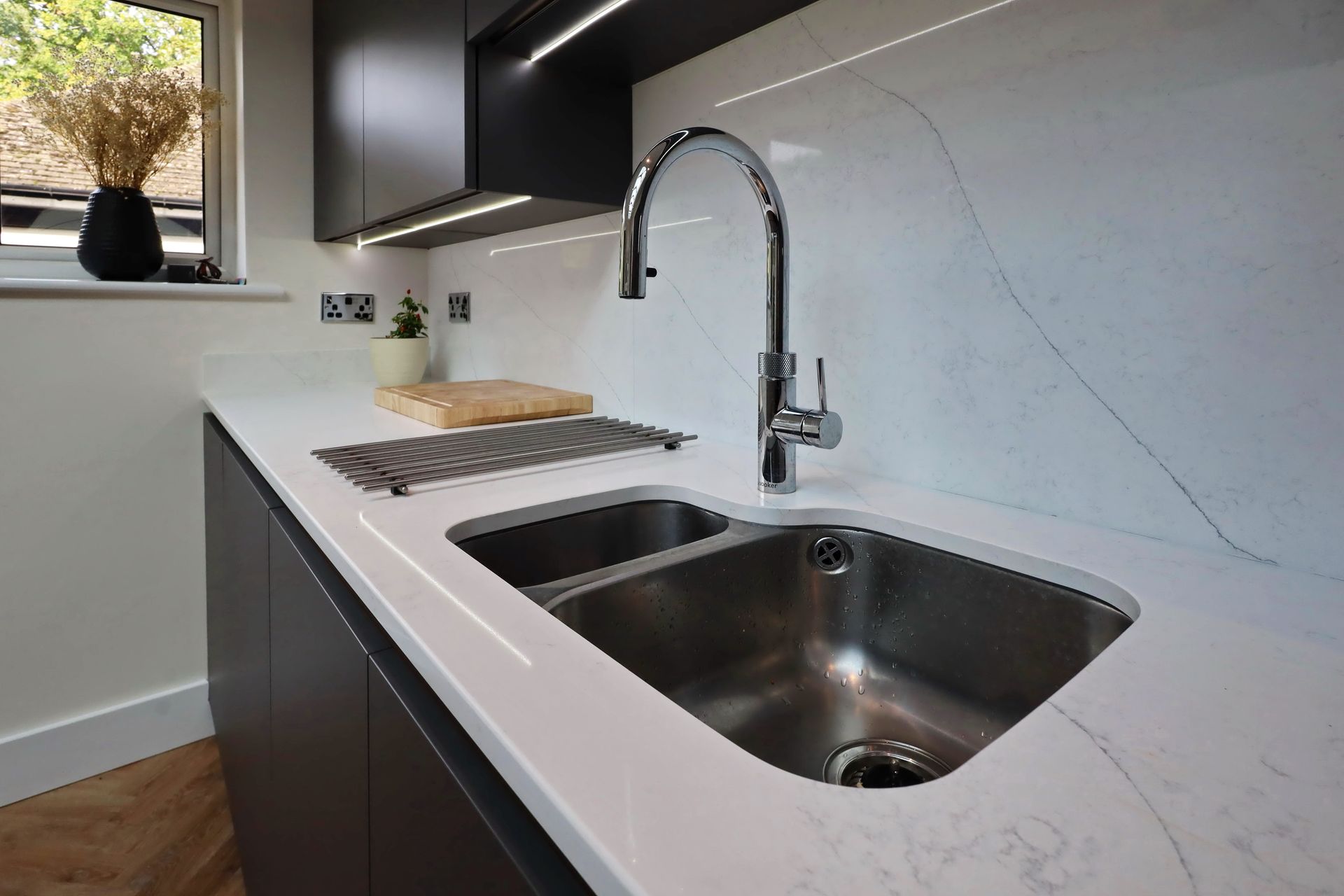 Modern kitchen sink in white countertop, stainless steel basin, with chrome faucet and dark cabinetry.