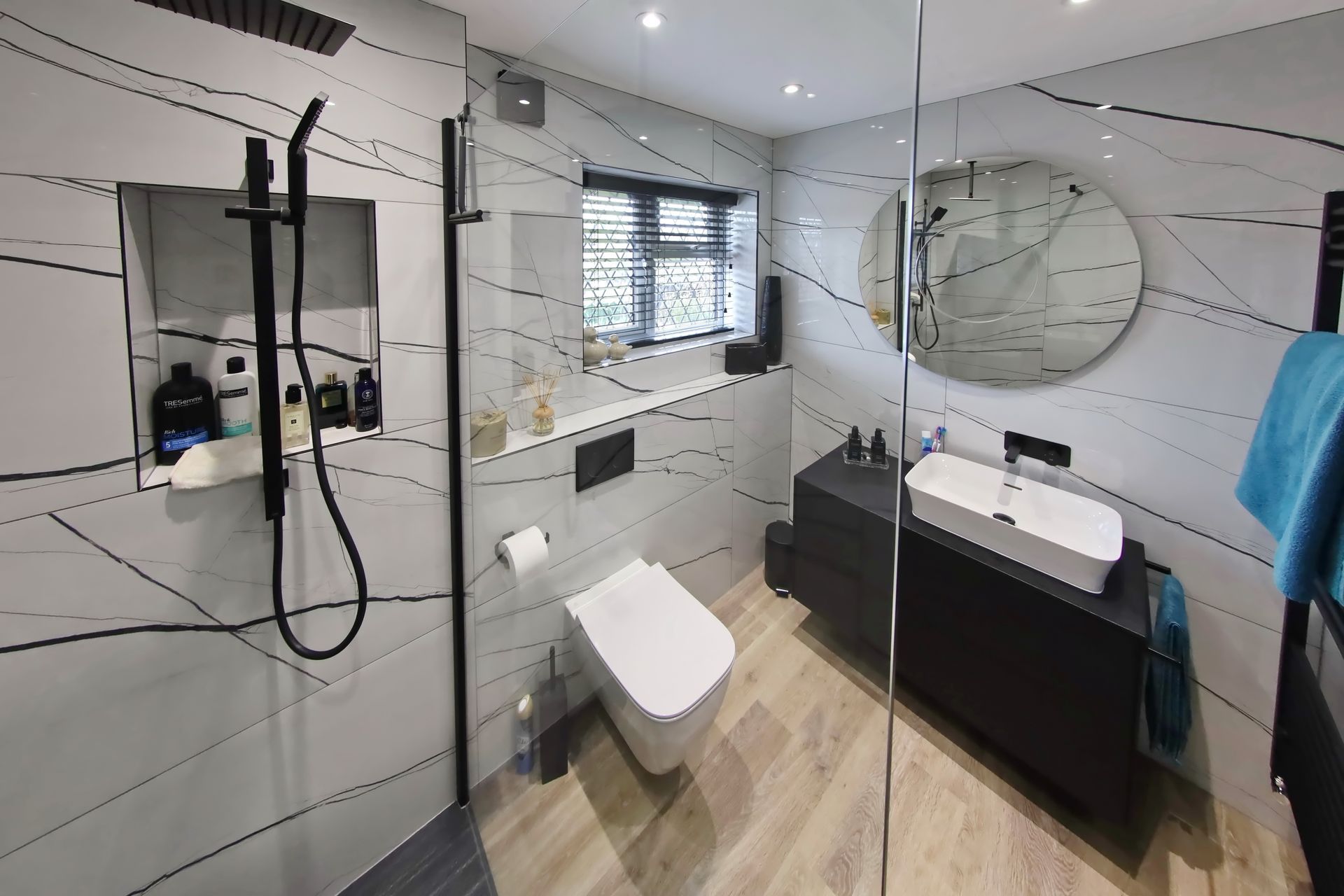 Modern bathroom with marble-look tile, black fixtures, glass shower, black vanity, and round mirror.