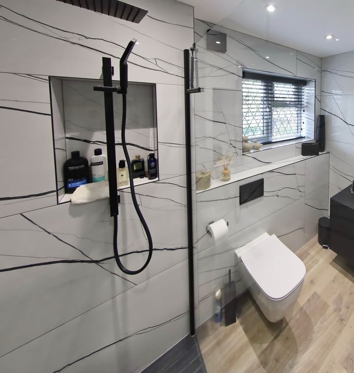 Modern bathroom with white and black marble-look tiles, black shower fixtures, toilet, and a window with blinds.