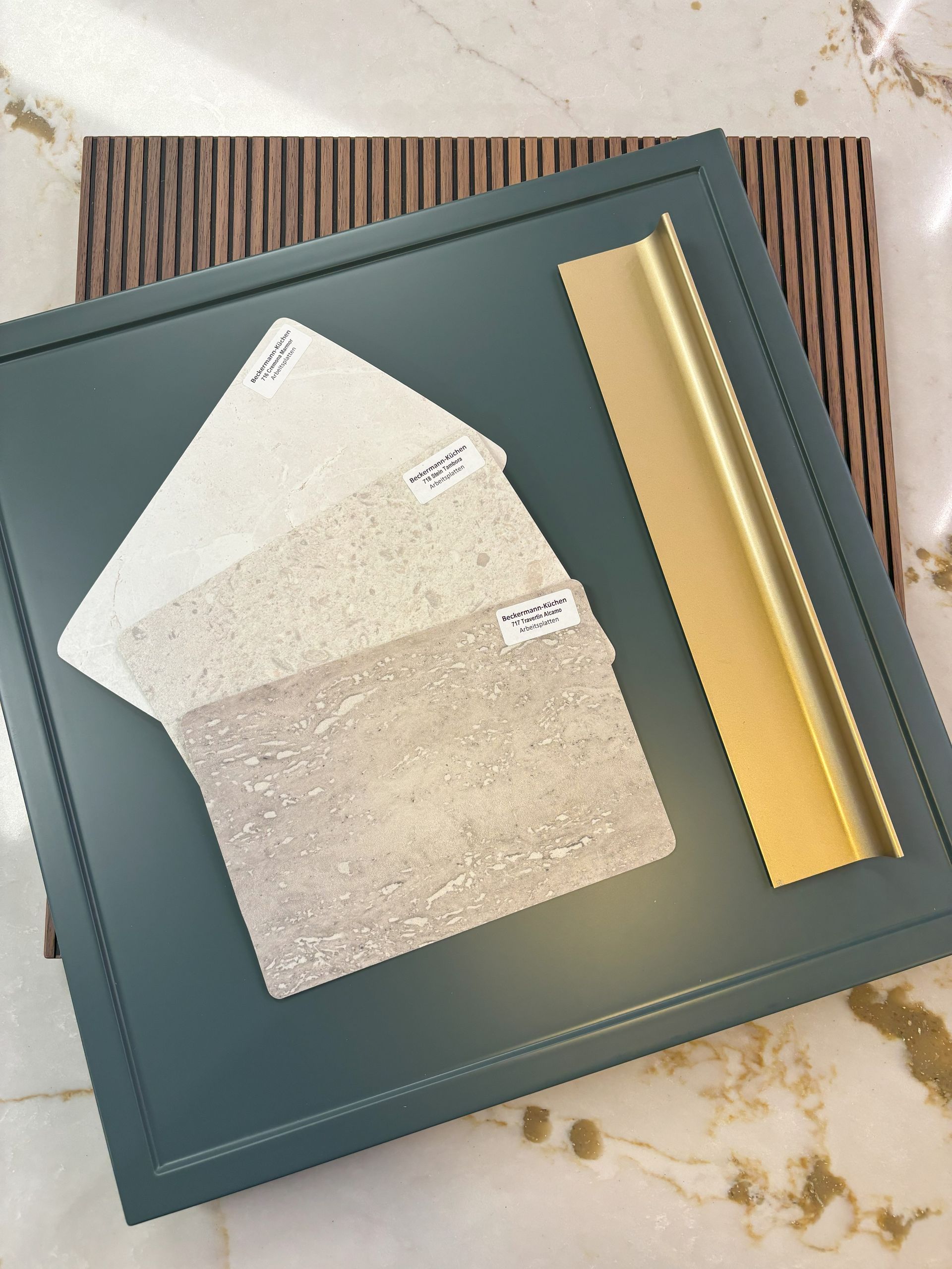 Mood board with blue-green cabinet door, beige stone samples, brass trim, and wood texture on a marble surface.