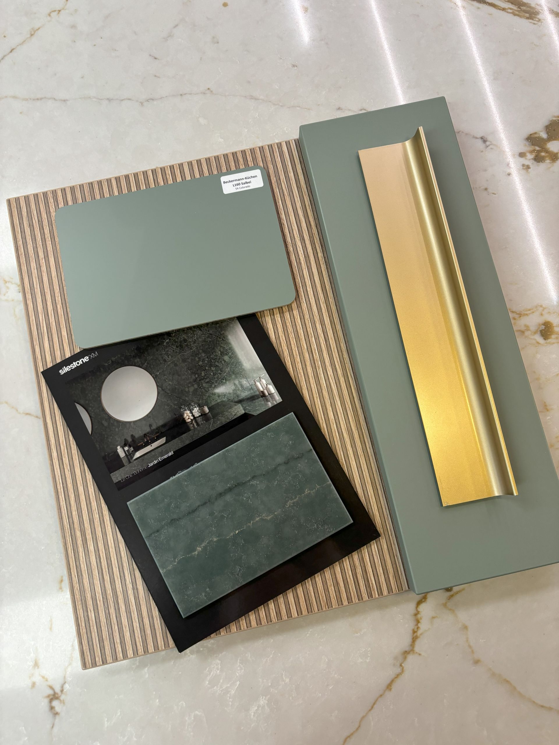 Color swatches and samples of interior design materials on a marble surface: green cabinet fronts, metal handle, and tile.