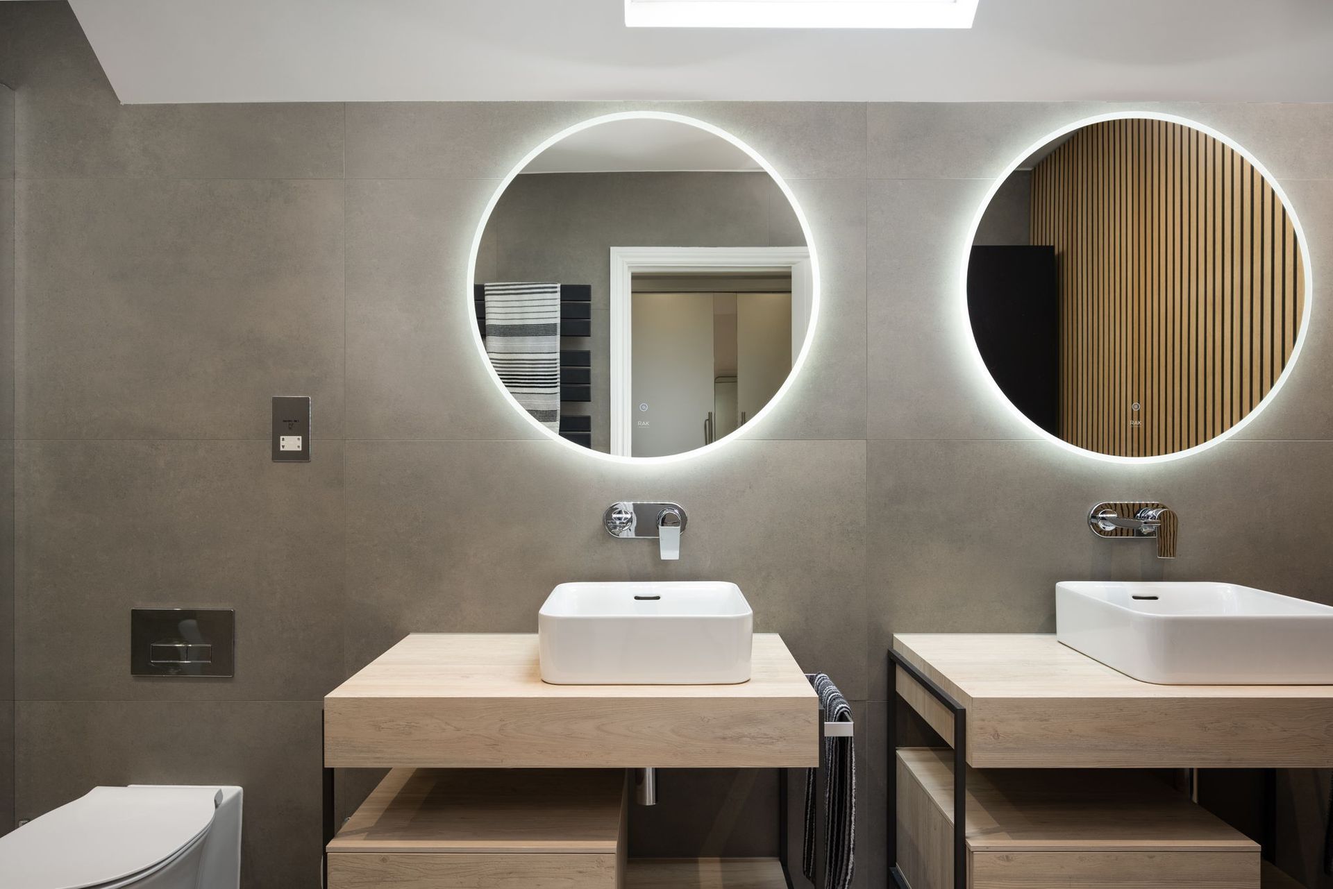 Modern bathroom with two circular mirrors with lights, floating wooden vanities, and grey tile.