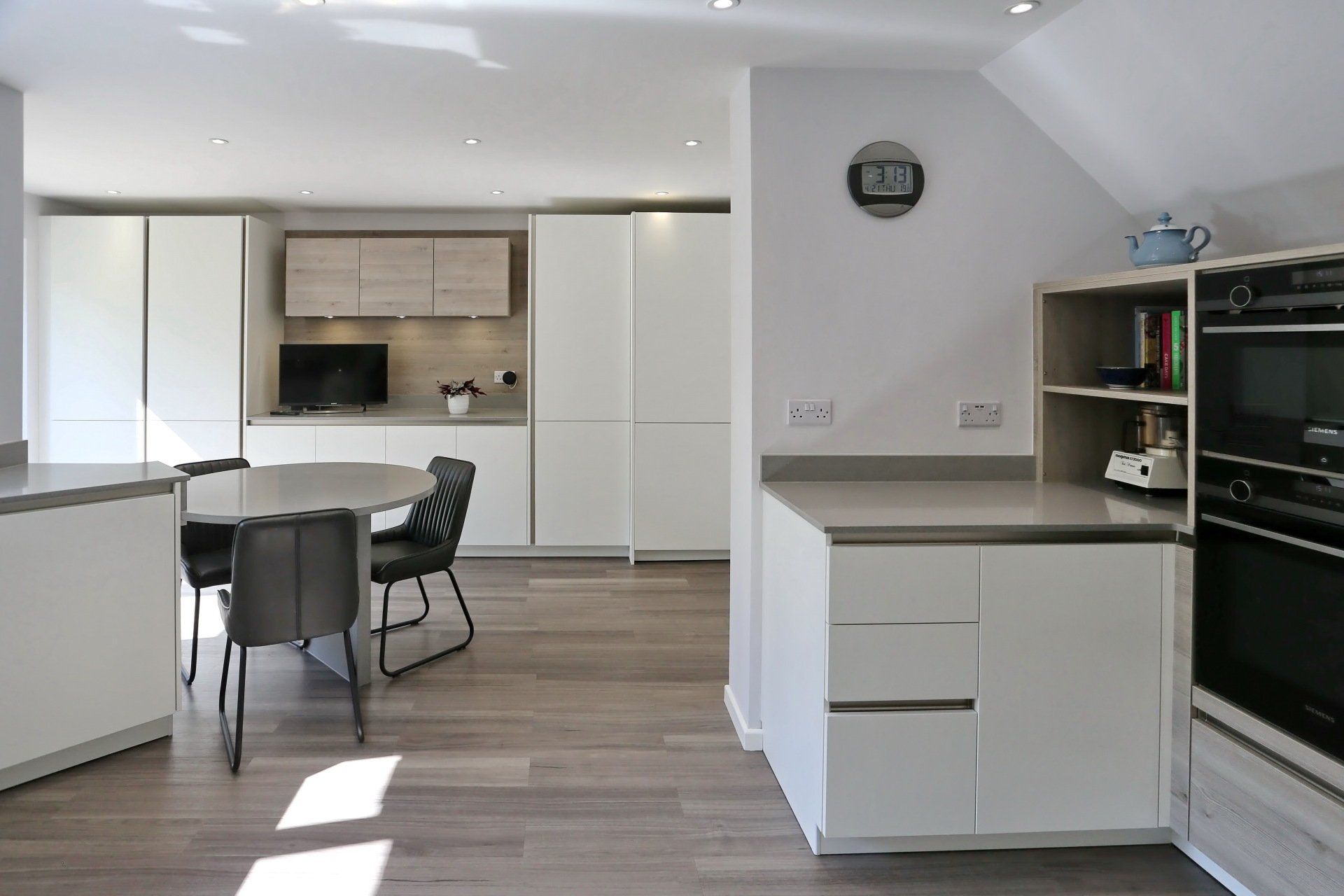 Modern kitchen with white cabinets, round table, grey countertops, and hardwood floors.