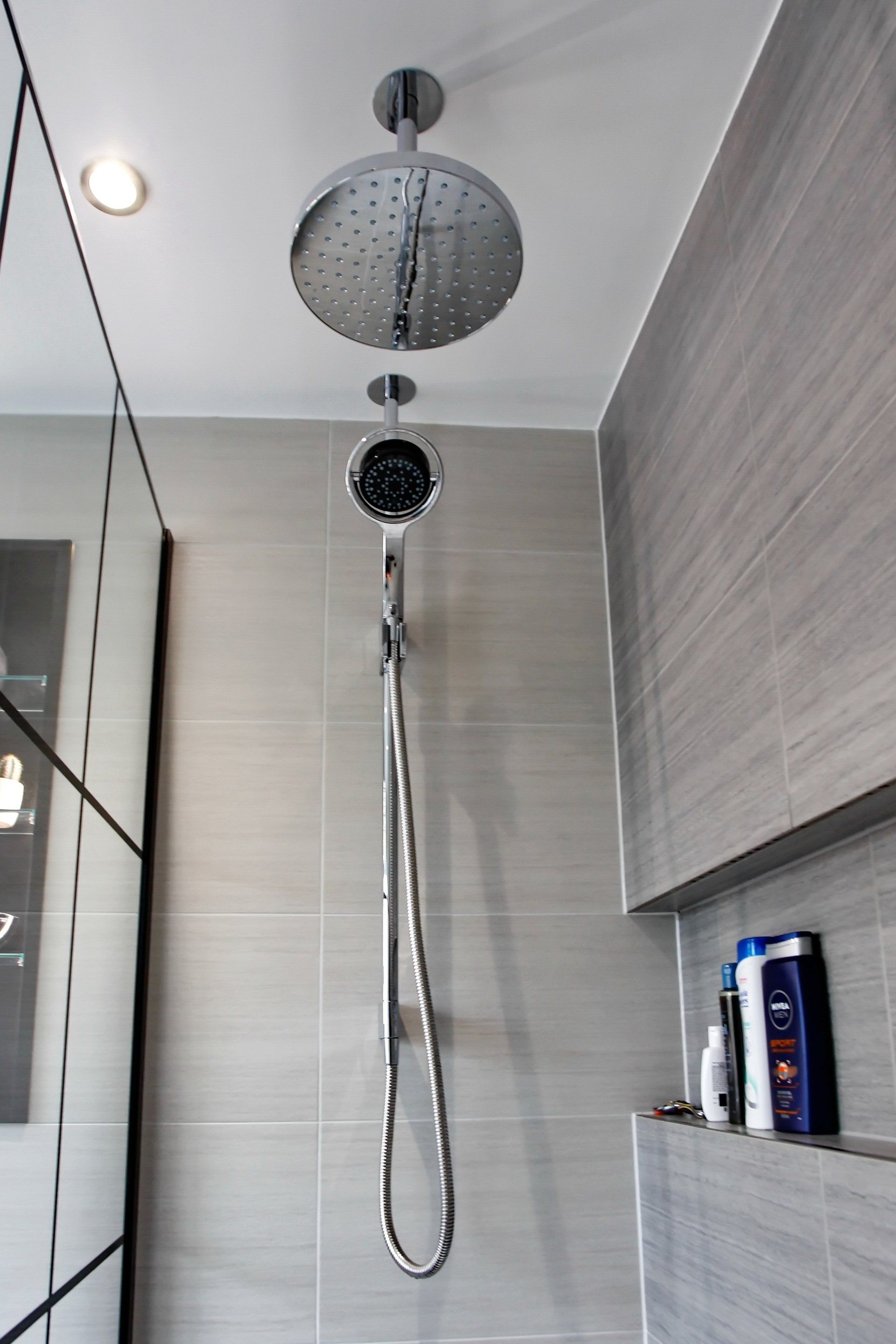 Chrome shower heads in a gray-tiled bathroom with a recessed shelf holding toiletries.
