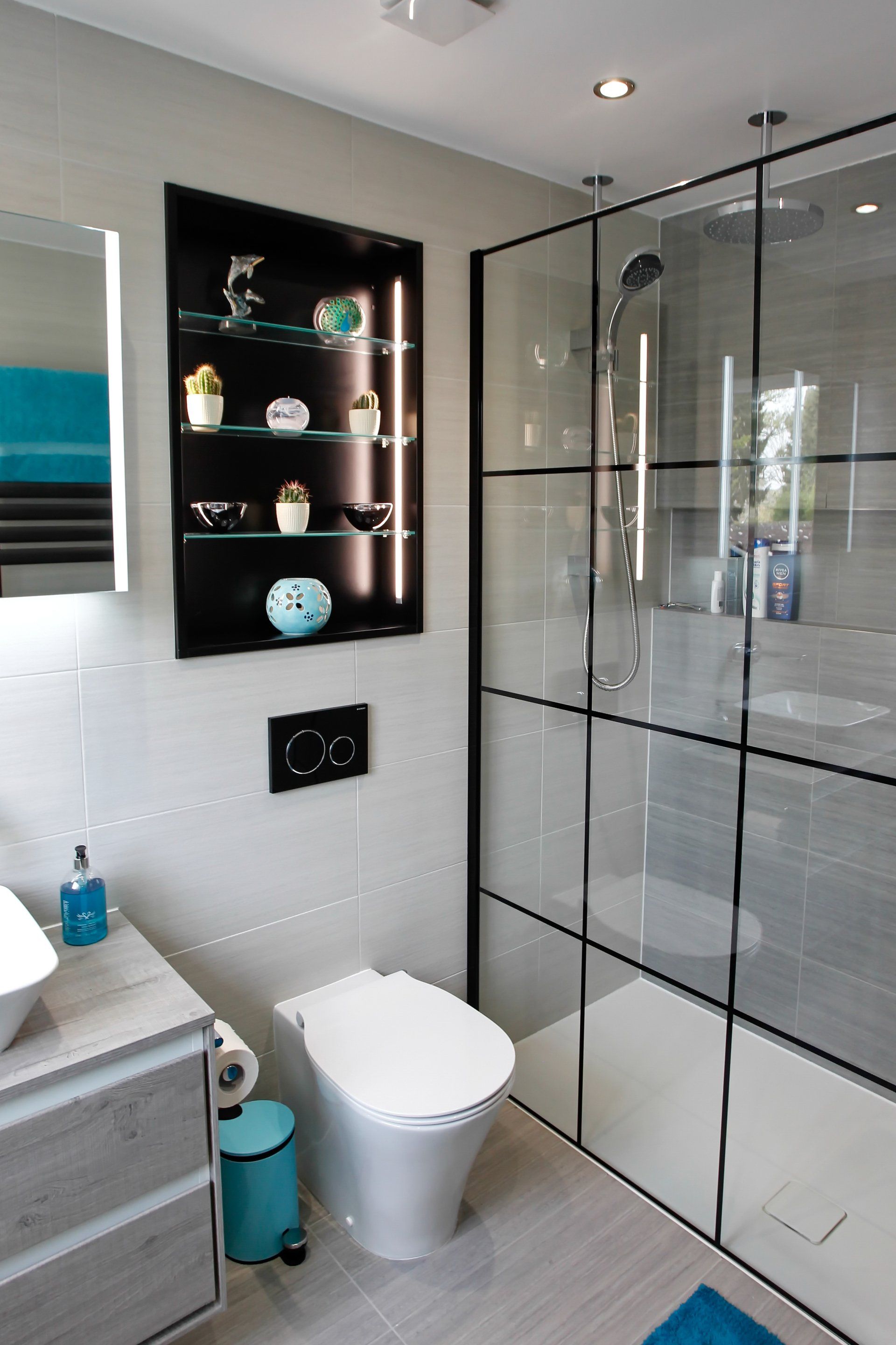 Modern bathroom with gray tiles, glass shower, black shelving, and a teal accent wall.