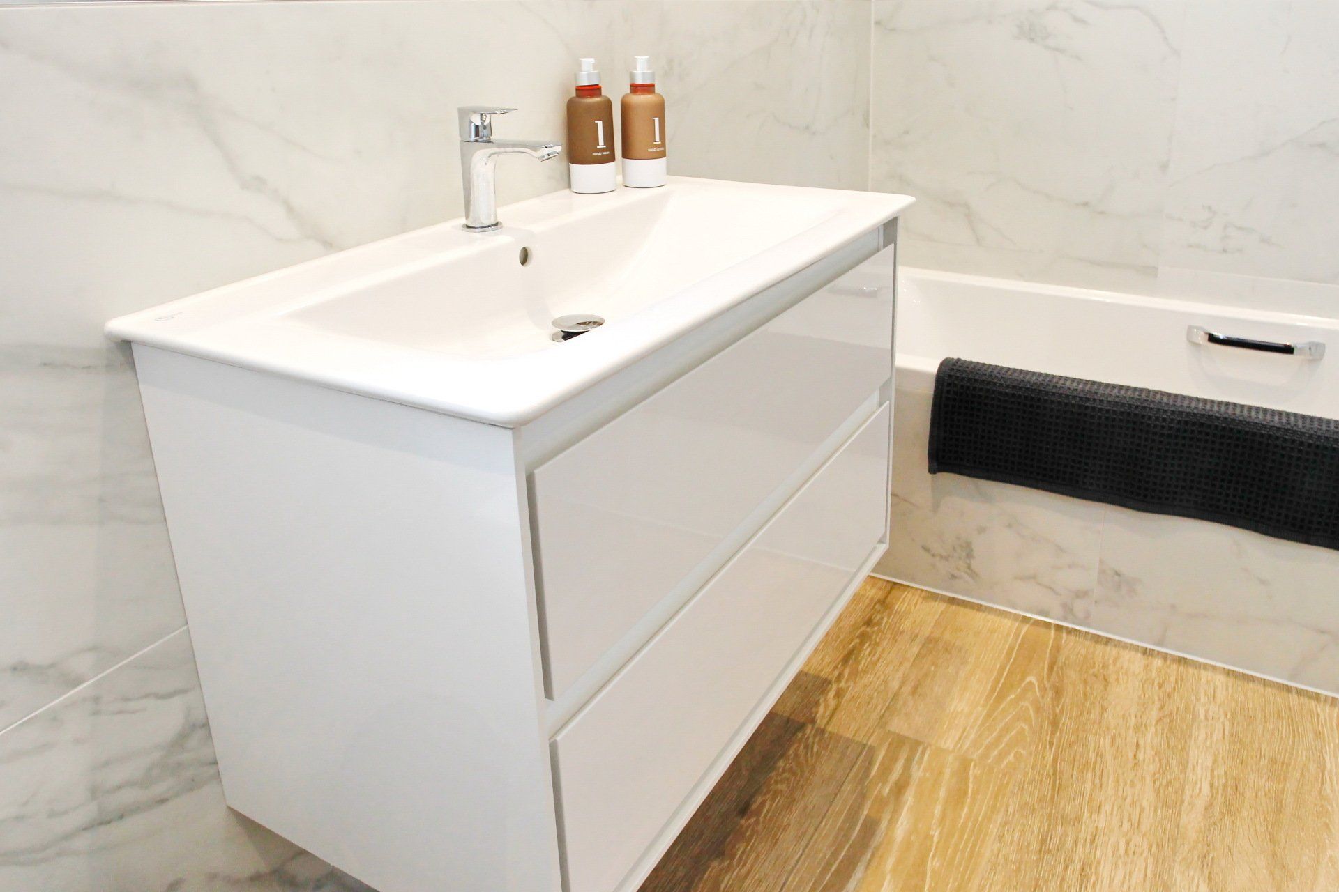 White vanity with two drawers and integrated sink in a bathroom, near a bathtub, with marble walls.