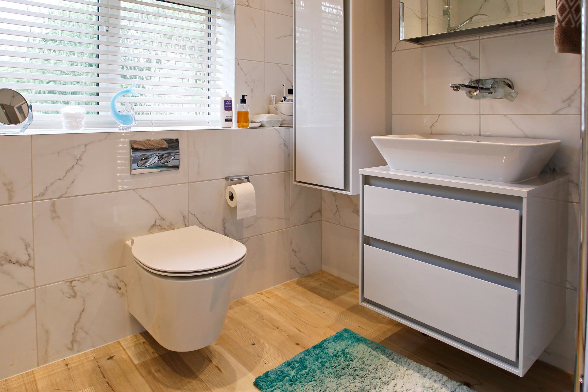 Modern white bathroom with floating toilet, vanity, and marble-look tiles. Light wood-look flooring and a window with blinds.