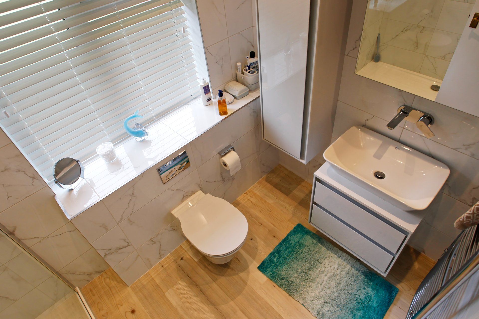Small modern bathroom with a toilet, sink, and storage cabinet; marble-effect walls, wooden floor, and a window with blinds.