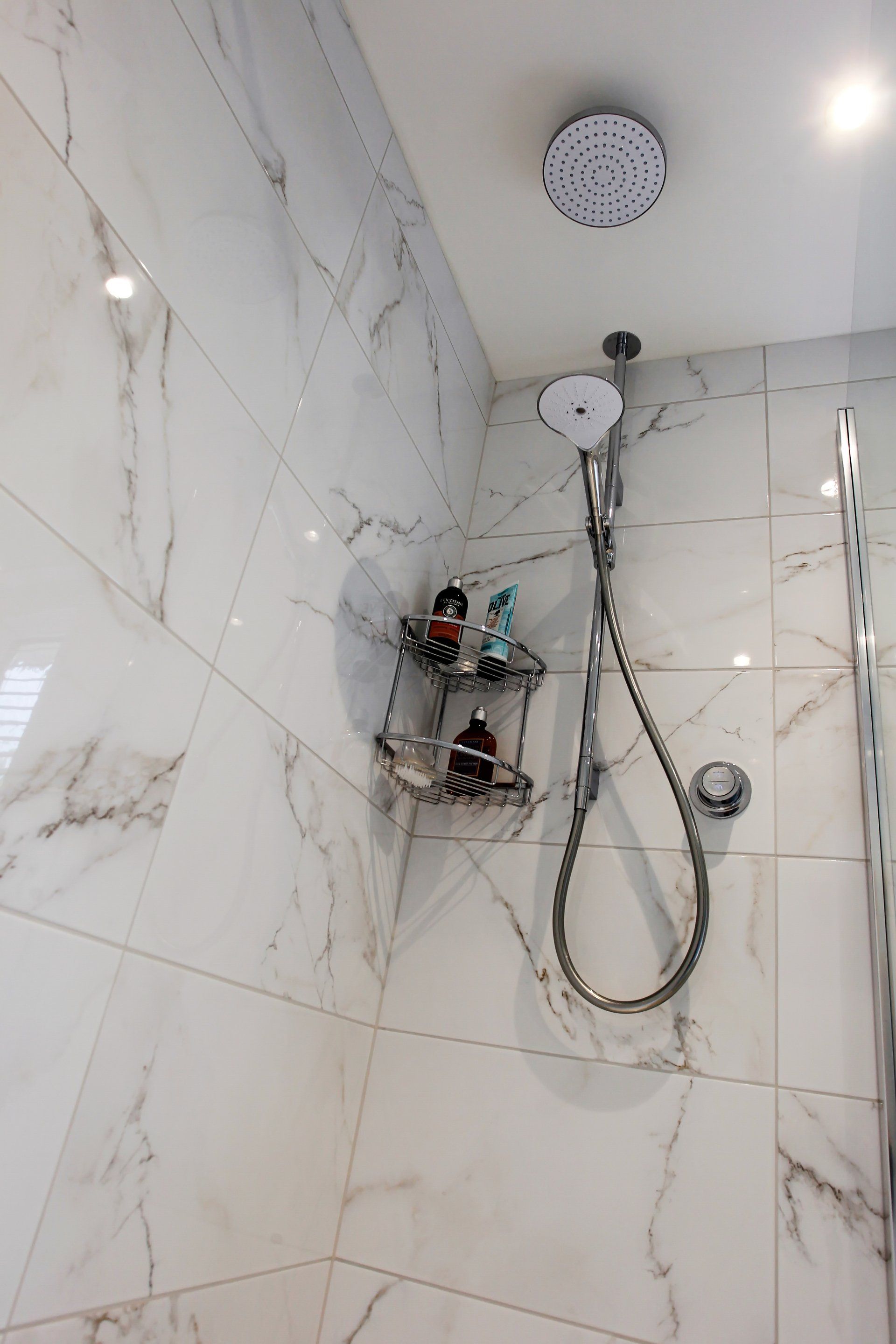 White marble-tiled shower with a rainfall shower head, handheld shower, and a shelf with toiletries.