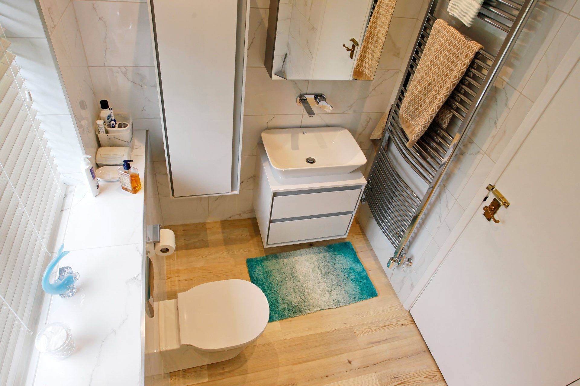 Small, modern bathroom with toilet, sink, and towel rack. Features white and wood tones with a teal rug.