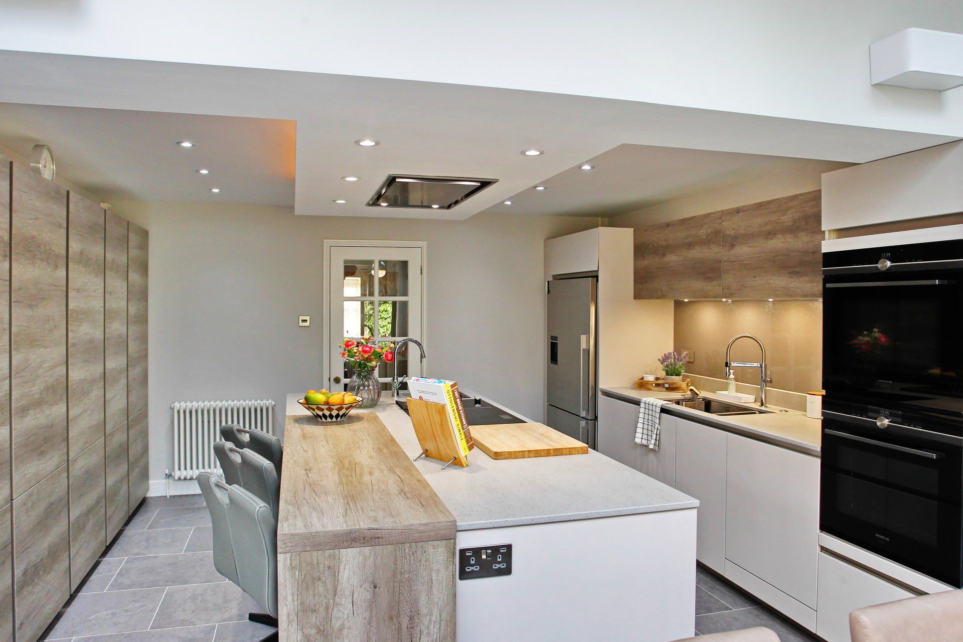 Modern kitchen with light wood accents, white cabinetry, island, built-in oven, and a window.