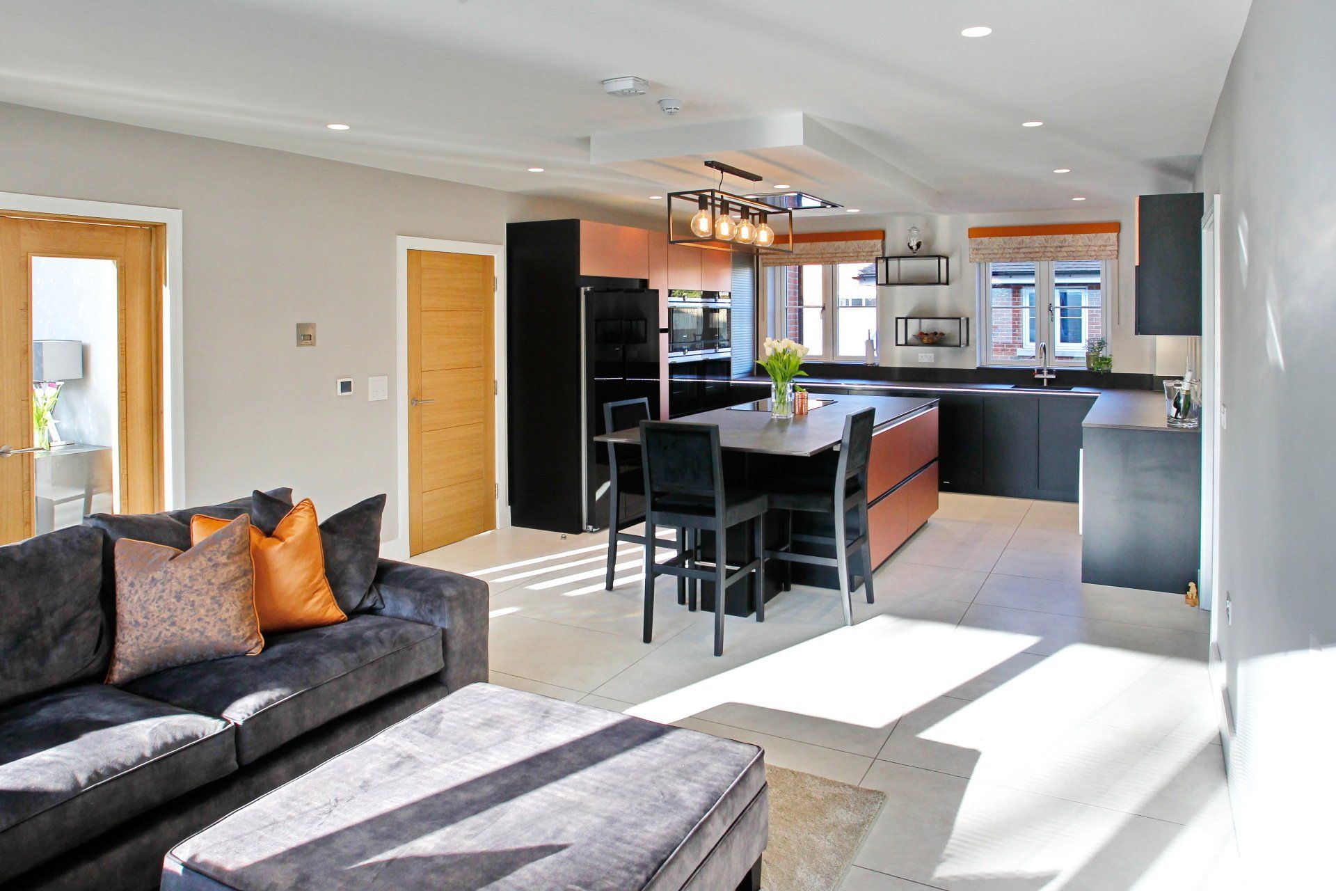 Open-plan kitchen/living room with dark cabinetry, orange accents, and a gray sofa. Sunlight streams in, illuminating the space.