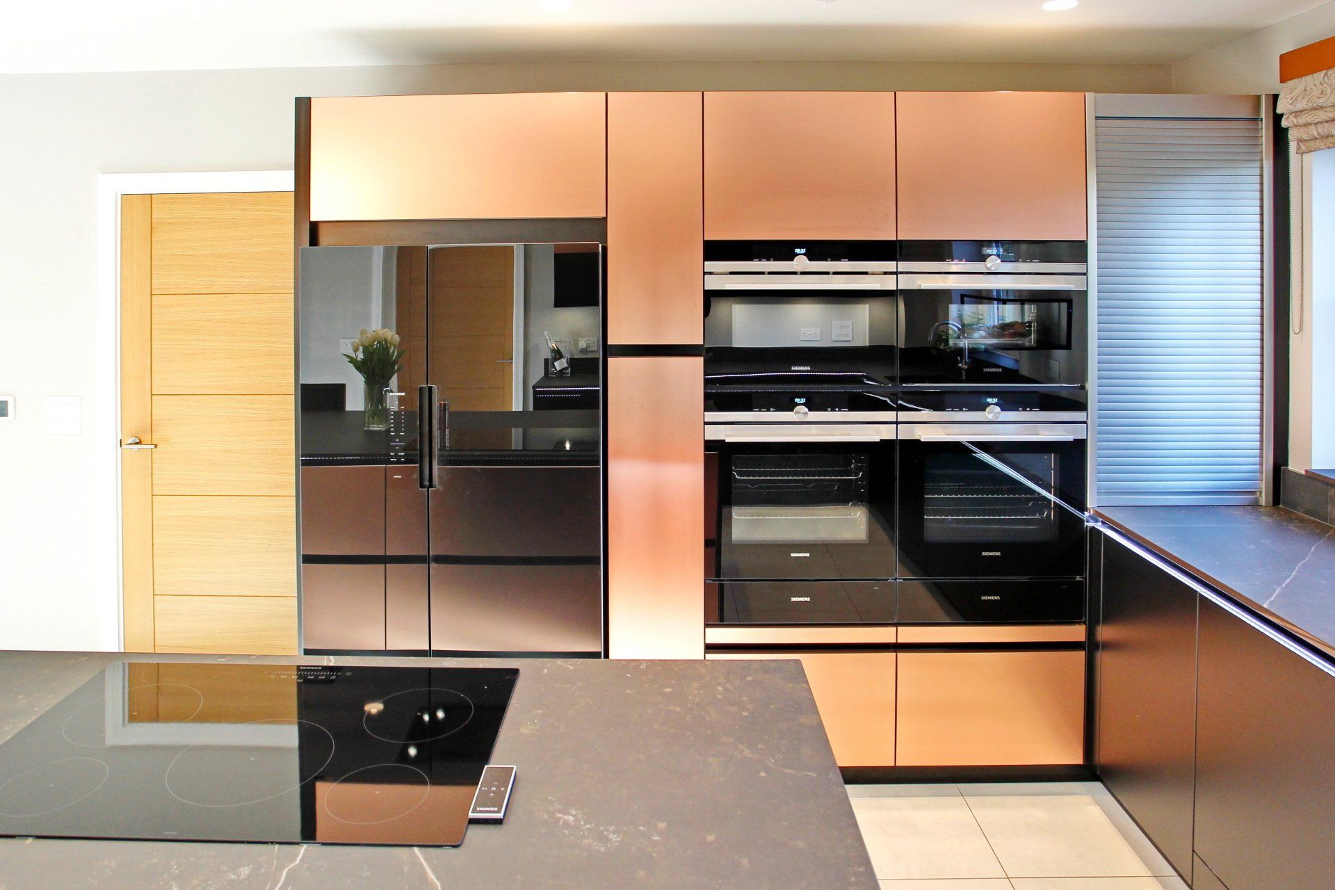 Copper-toned kitchen cabinets and appliances; refrigerator and ovens are visible. Dark countertop in foreground.