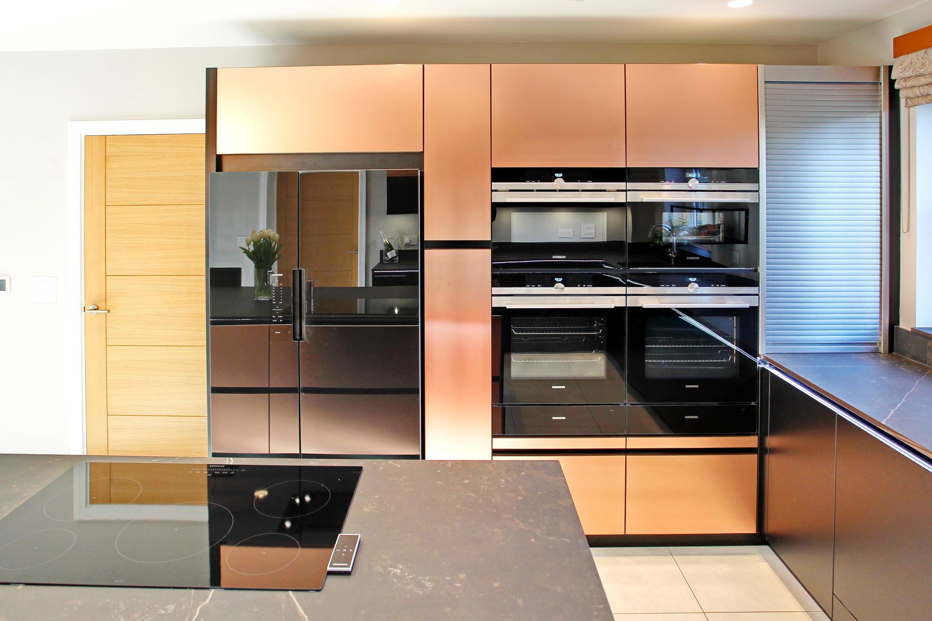 Copper-colored kitchen cabinets and appliances with a black countertop and a light wood door.