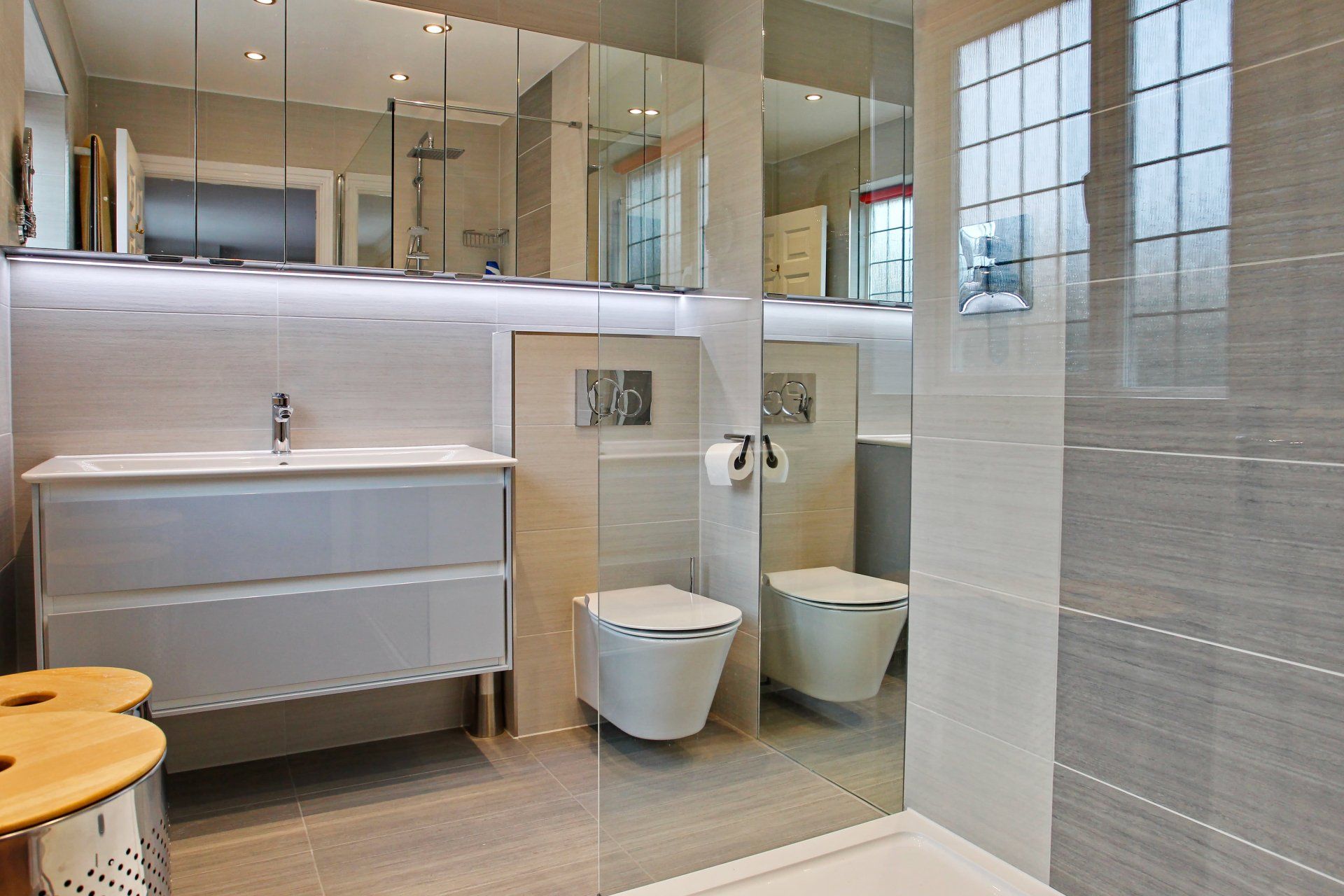 Modern bathroom with gray and beige tiles, floating toilet and sink, large mirrors, and shower.