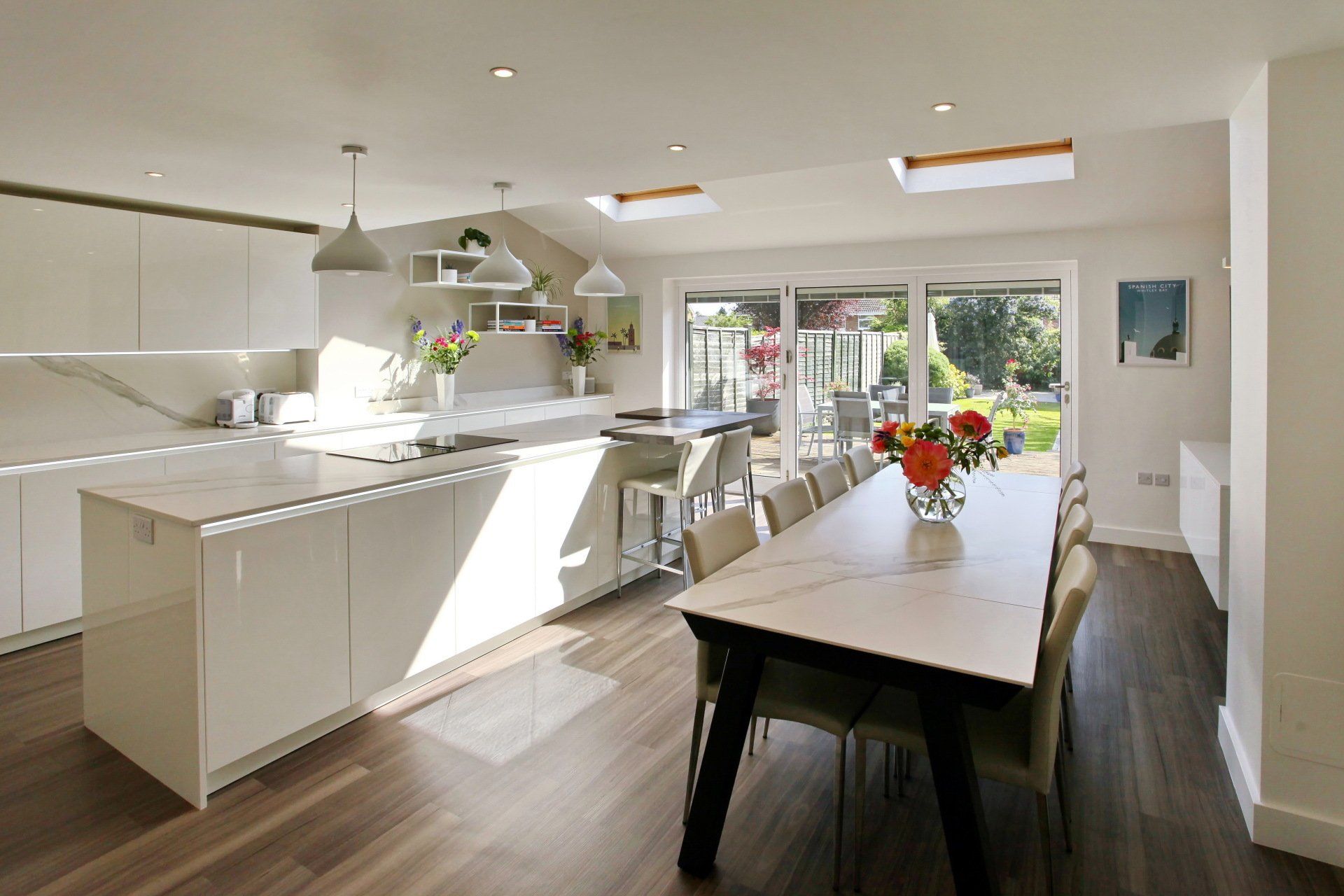 Modern white kitchen with island, marble countertops, dining table, and large windows overlooking a backyard.