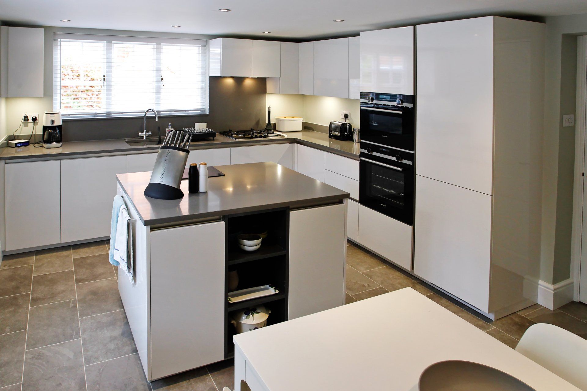 Modern white kitchen with island and integrated appliances; gray countertops and tiled floor.