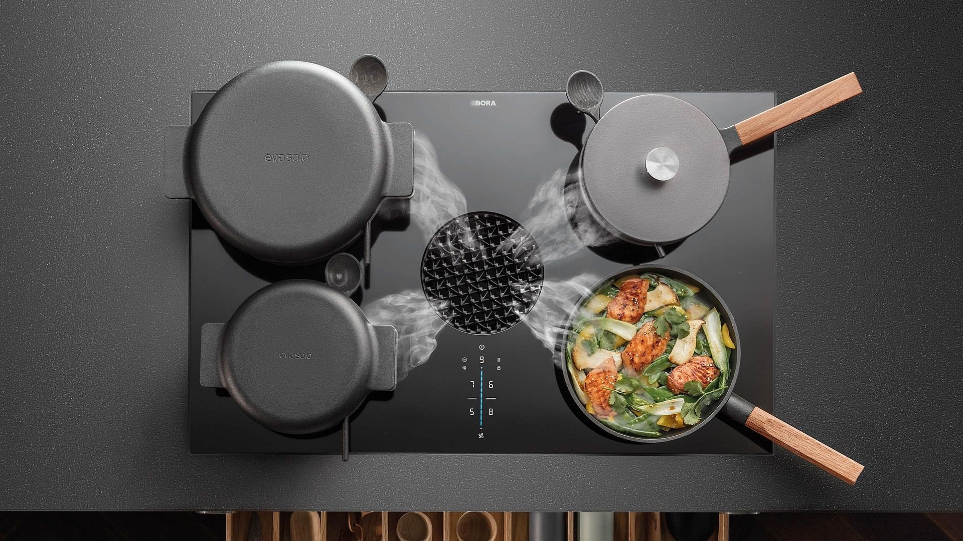 A black cooktop with four pots/pans cooking and steam rising around a central vent.